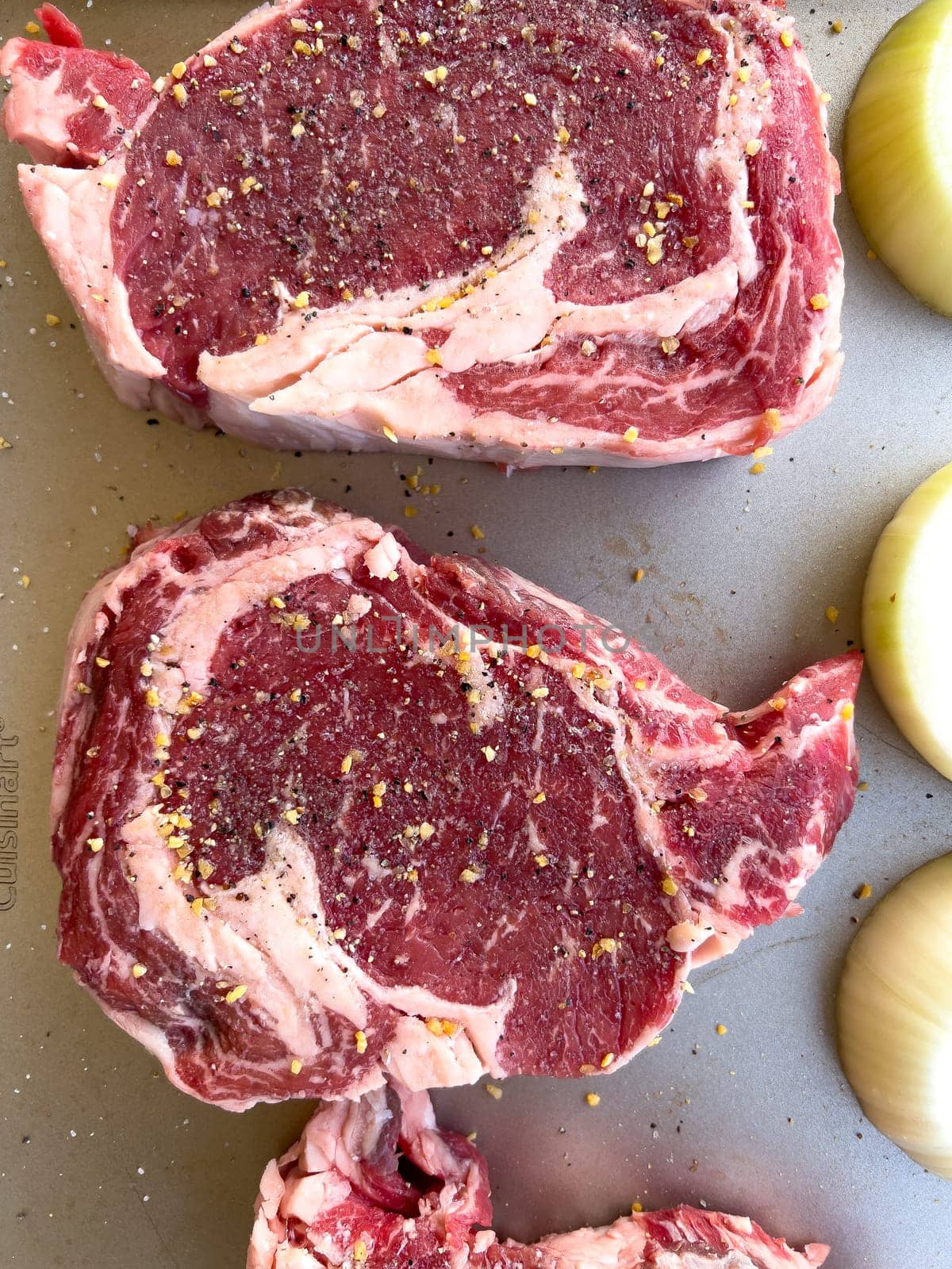 Three raw ribeye steaks with marbling, liberally seasoned with coarse spices, lie beside cut halves of onions on a kitchen tray, ready for cooking.