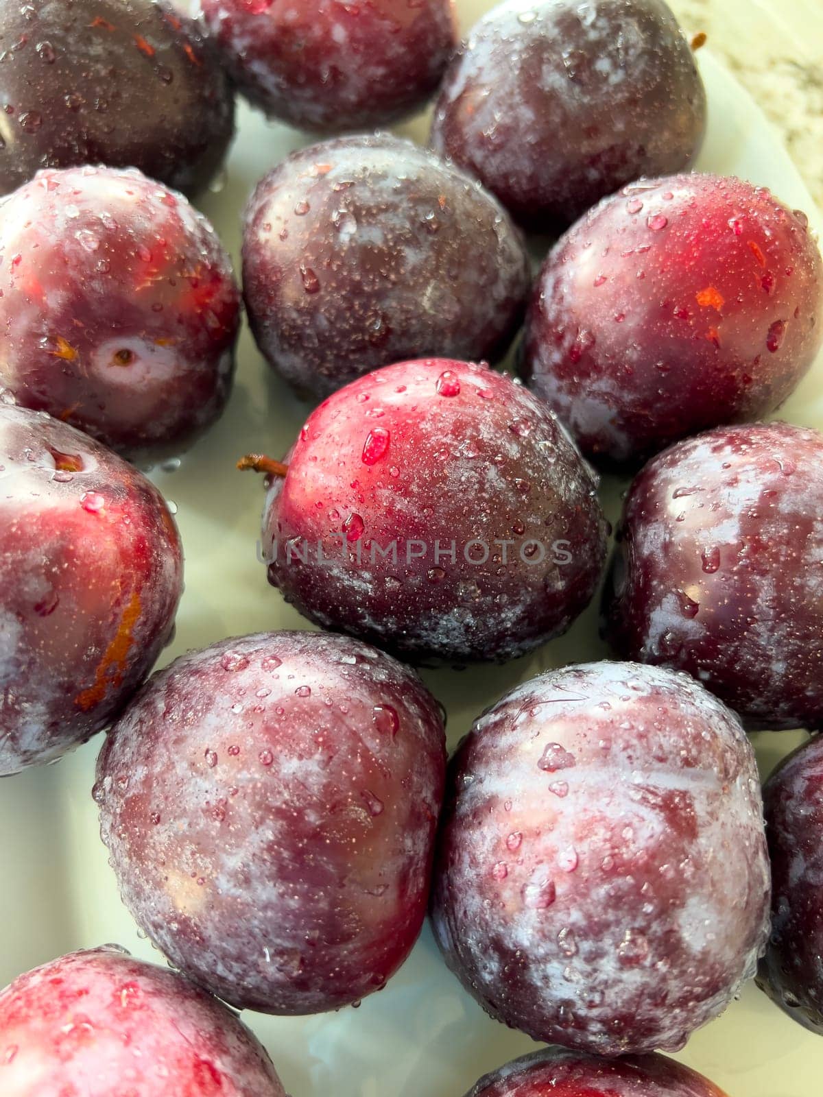 A plate of ripe, frosty plums sits prominently on a marble countertop in a sunlit kitchen with a crisp, white interior and neatly arranged appliances.