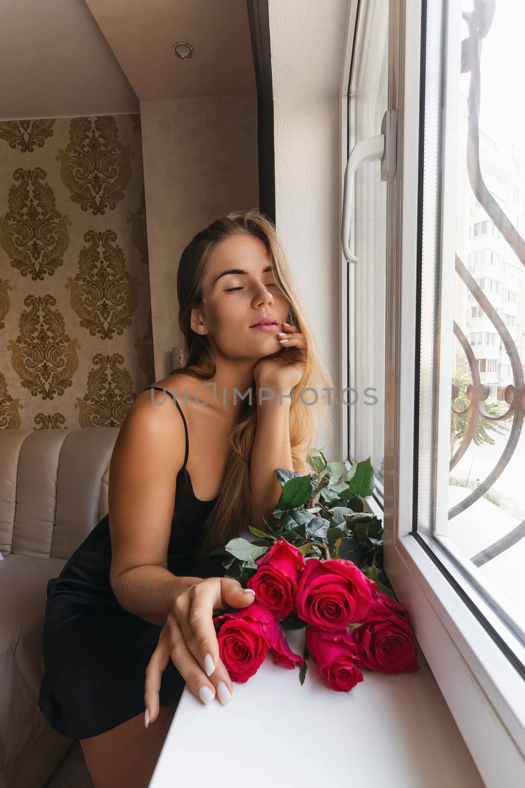 A woman in a black dress is sitting in front of a window with a bouquet of red roses