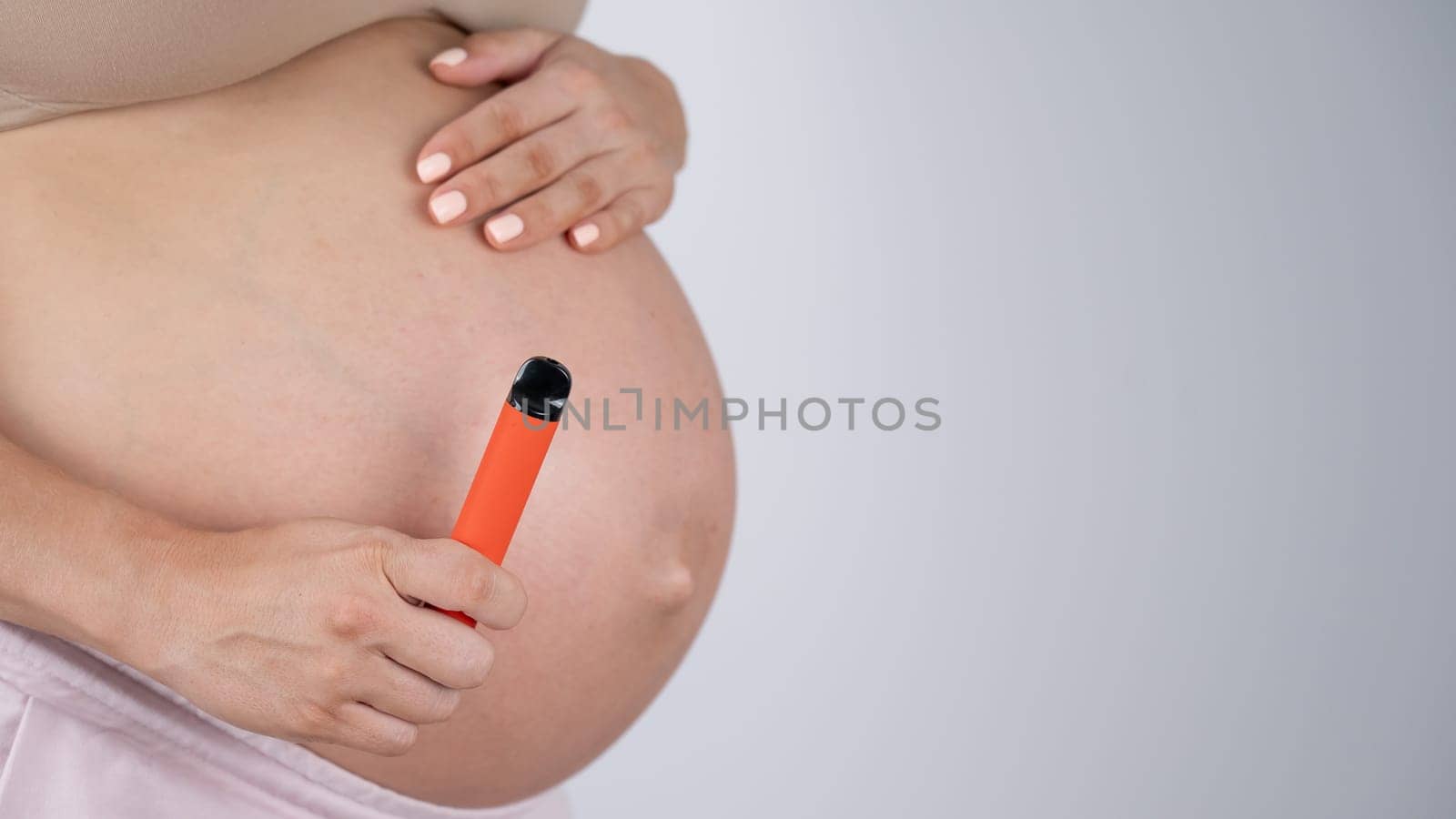 A pregnant woman smokes a vape. A girl holds an electronic cigarette against the background of her bare tummy. Copy space