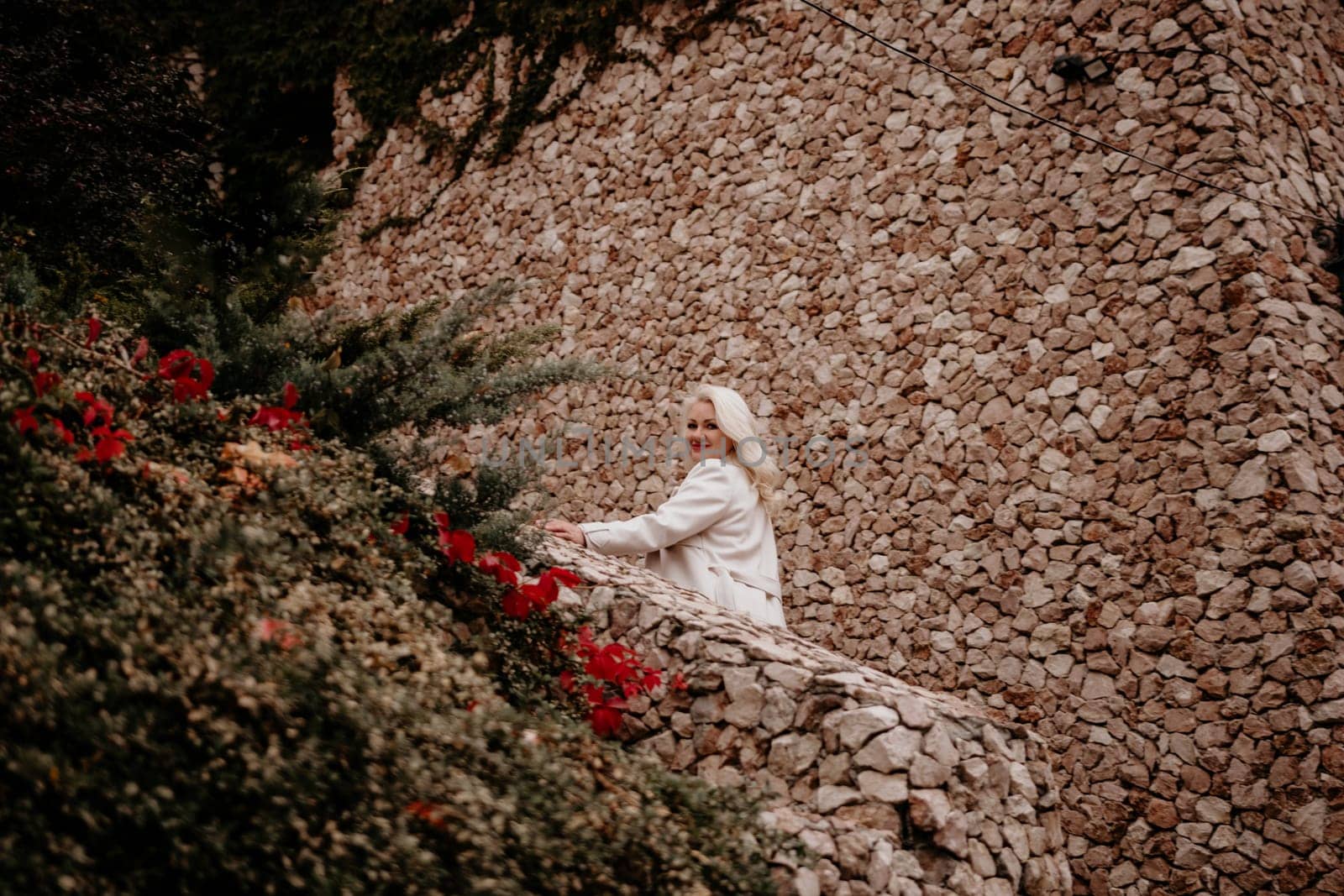 A woman is sitting on a stone wall with red flowers in the background. She is smiling and looking at the camera