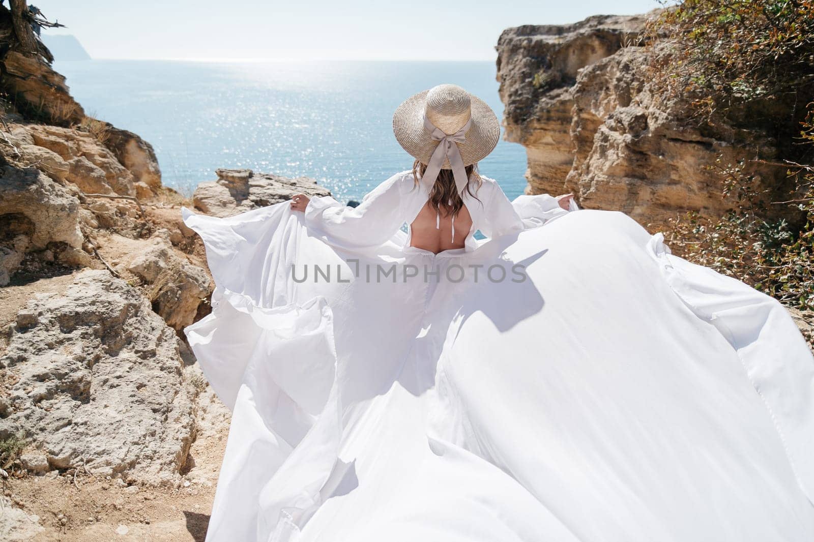 A woman in a white dress is standing on a rocky beach with her hat on. The scene is serene and peaceful, with the woman enjoying the view of the water by Matiunina