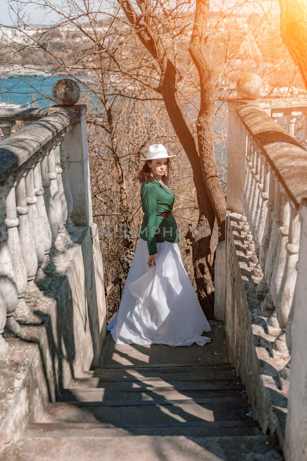 Woman walks around the city, lifestyle. Happy woman in a green jacket, white skirt and hat is sitting on a white fence with balusters overlooking the sea bay and the city. by Matiunina