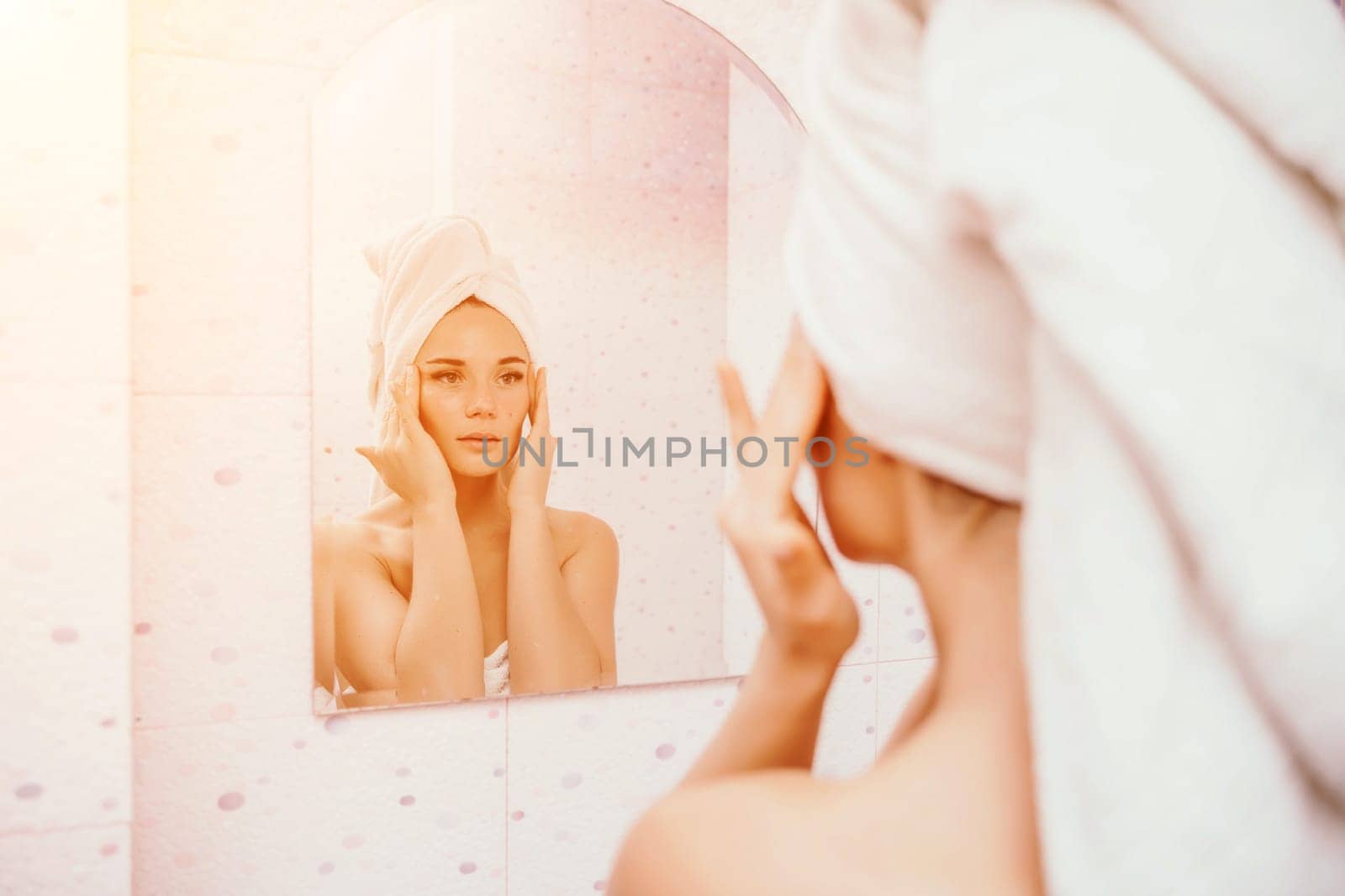 Young beautiful woman using face cream moisturizing lotion after bath. Pretty attractive girl in a towel on her head stands in front of a mirror in a home bathroom. Daily hygiene and skin care by Matiunina