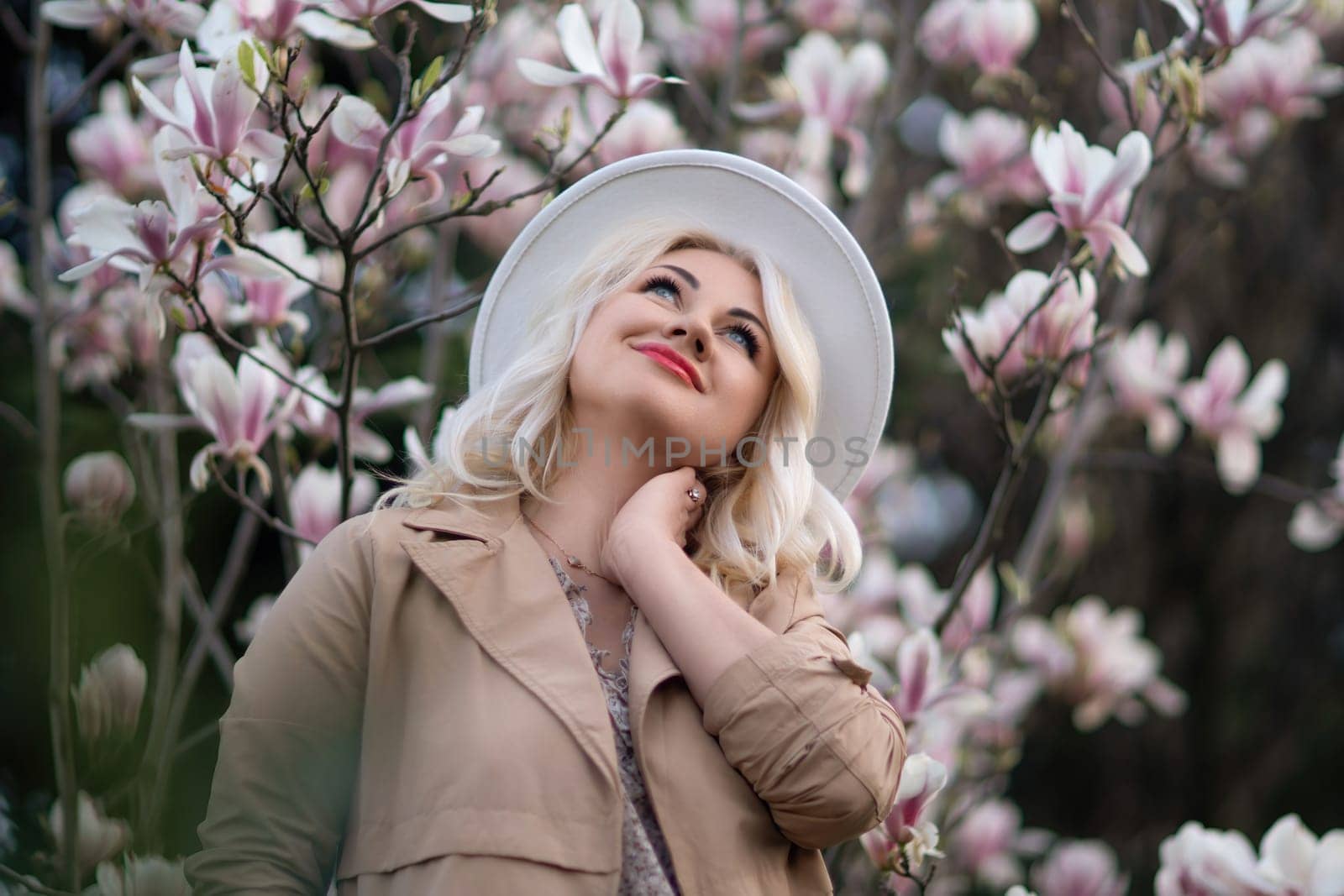 Magnolia flowers woman. A blonde woman wearing a white hat stands in front of a tree full of pink flowers. She is wearing a brown jacket and a necklace