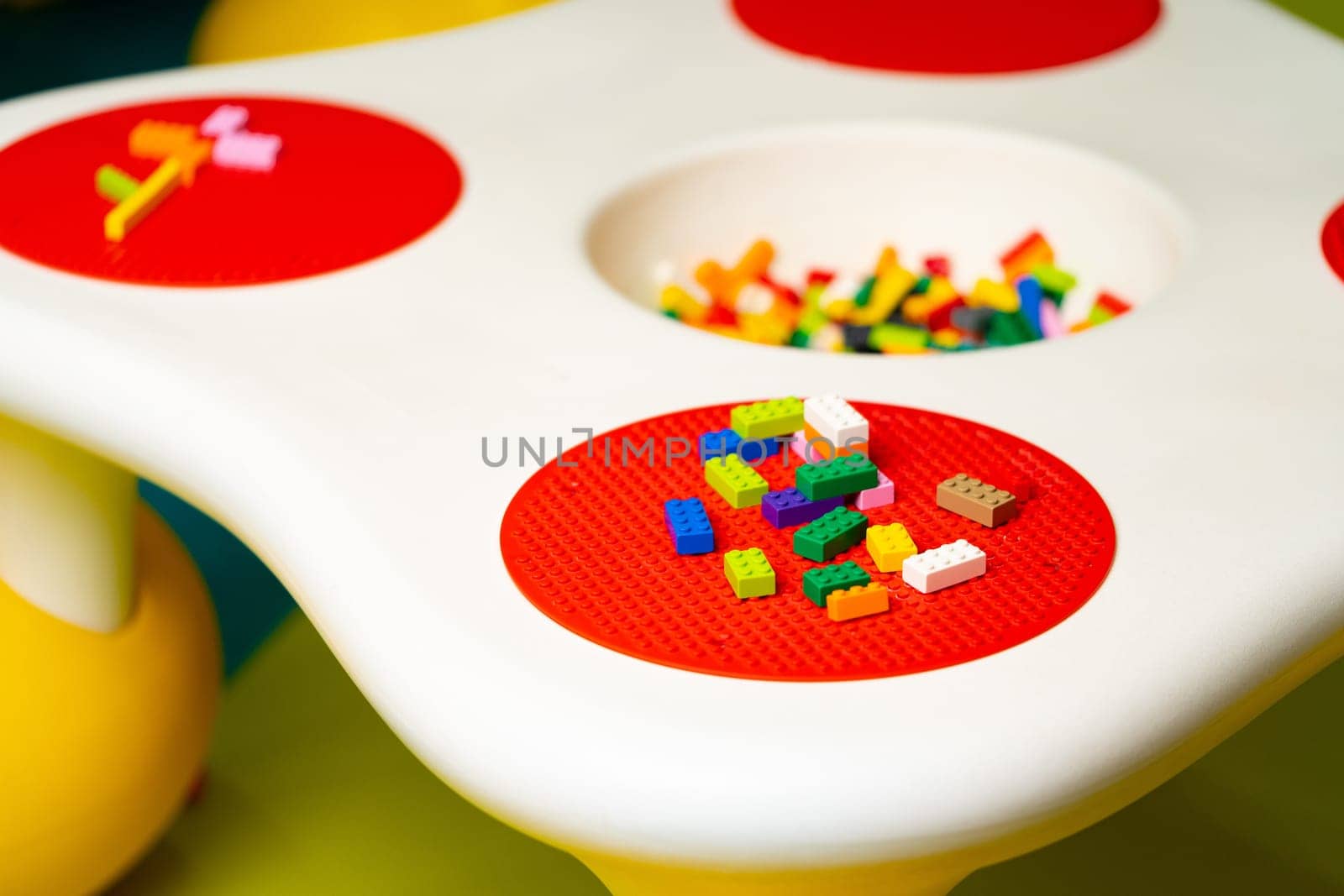 A close-up view of a childs play table filled with colorful toys. Various toys like blocks, dolls, cars, and puzzles are scattered around the table. The childs hands are seen playing and interacting with the toys.