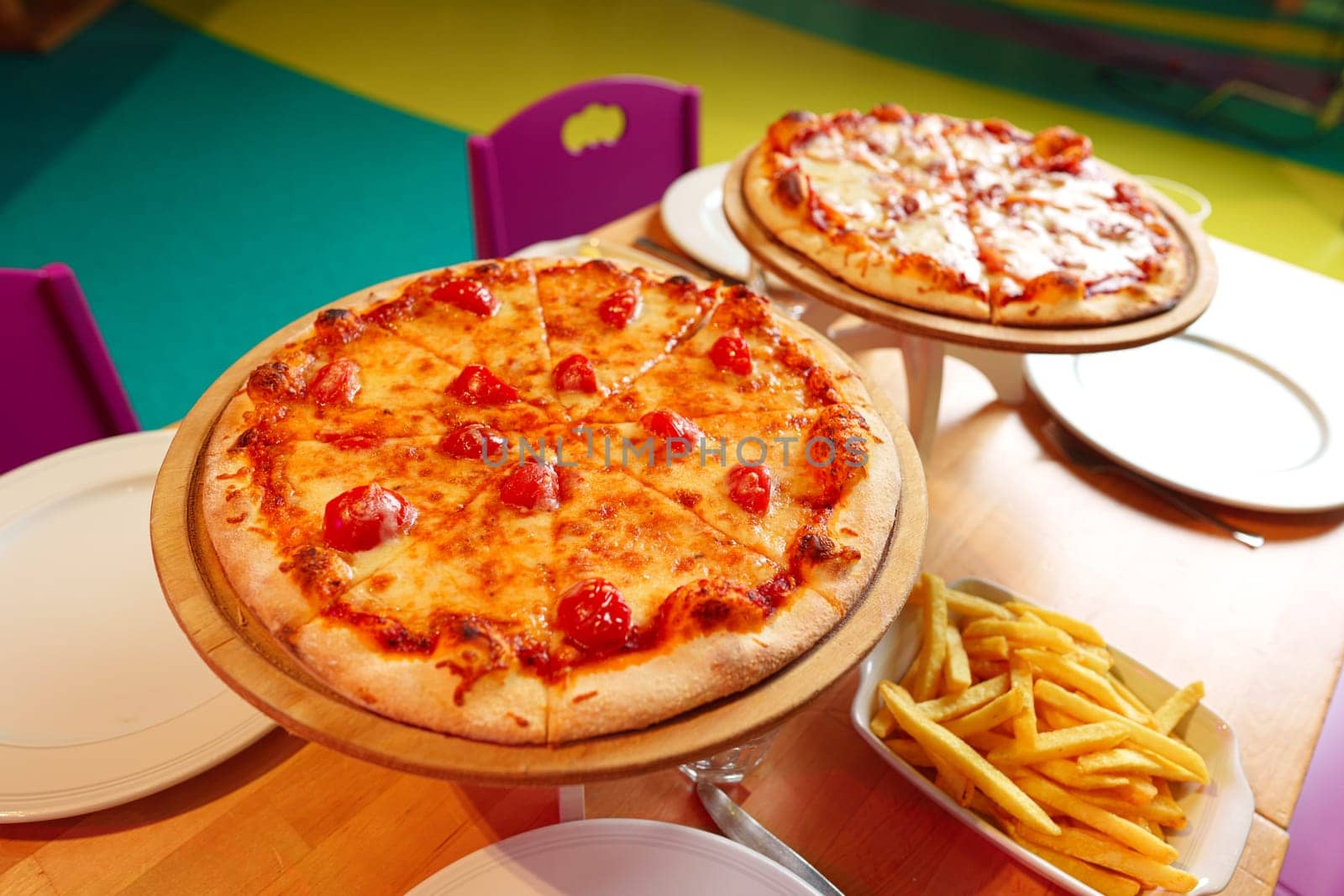 Two delicious pizzas with golden-brown cheese are displayed on elevated wooden stands above a table adorned with a colorful tablecloth. Beside the pizzas, there is a bowl of golden French fries and glasses filled with amber-colored beverages, suggesting an informal outdoor meal.