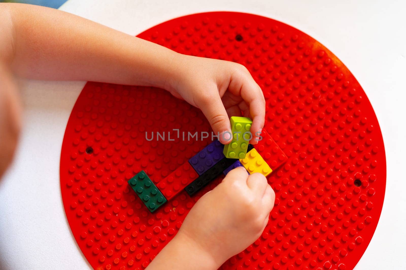 A young child is deeply focused while constructing a design with colorful plastic building blocks on a round red base. The childs hands are precisely placing a blue block to continue the pattern they have started, indicating an exercise in fine motor skills and creativity.