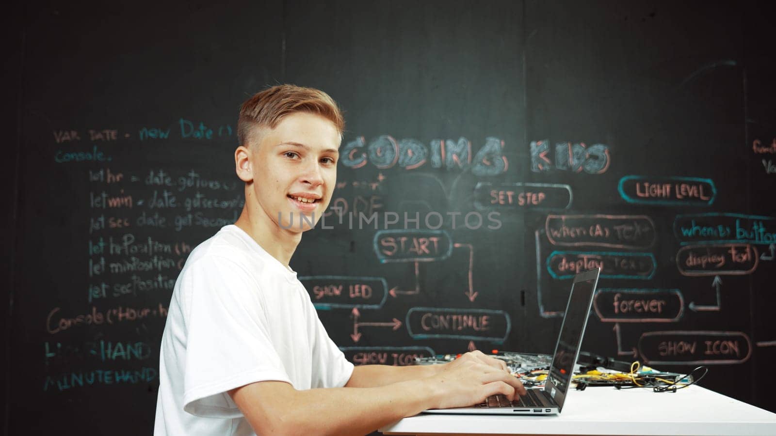 Caucasian boy coding and programing system while looking at laptop screen and camera with confident. Attractive academic student using computer analysis data while sitting at blackboard. Edification.