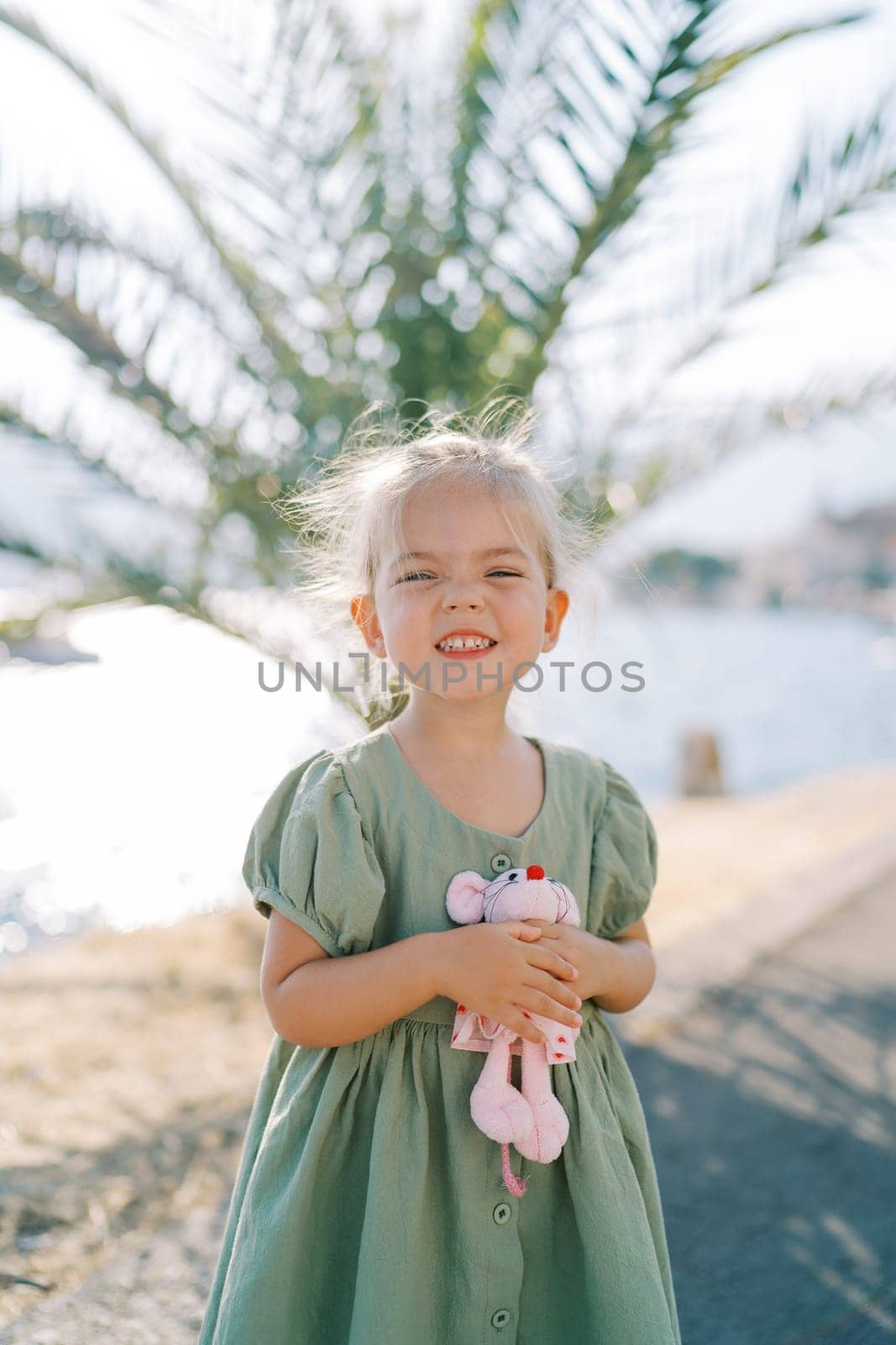 Little smiling girl with a toy pink mouse in her hand stands on the seashore near a green palm tree. High quality photo