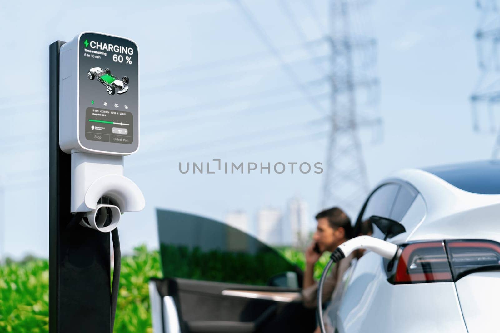 Man talking on the phone while recharge EV car battery at charging station connected to power grid tower electrical as electrical industry for eco friendly car utilization.Expedient