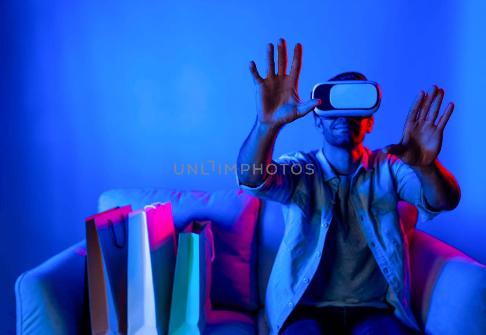 Caucasian man with vr glass posing pointing finger while sitting at sofa with shopping bags. Person selecting and doing shopping online by using VR technology and virtual goggle at home. Deviation.