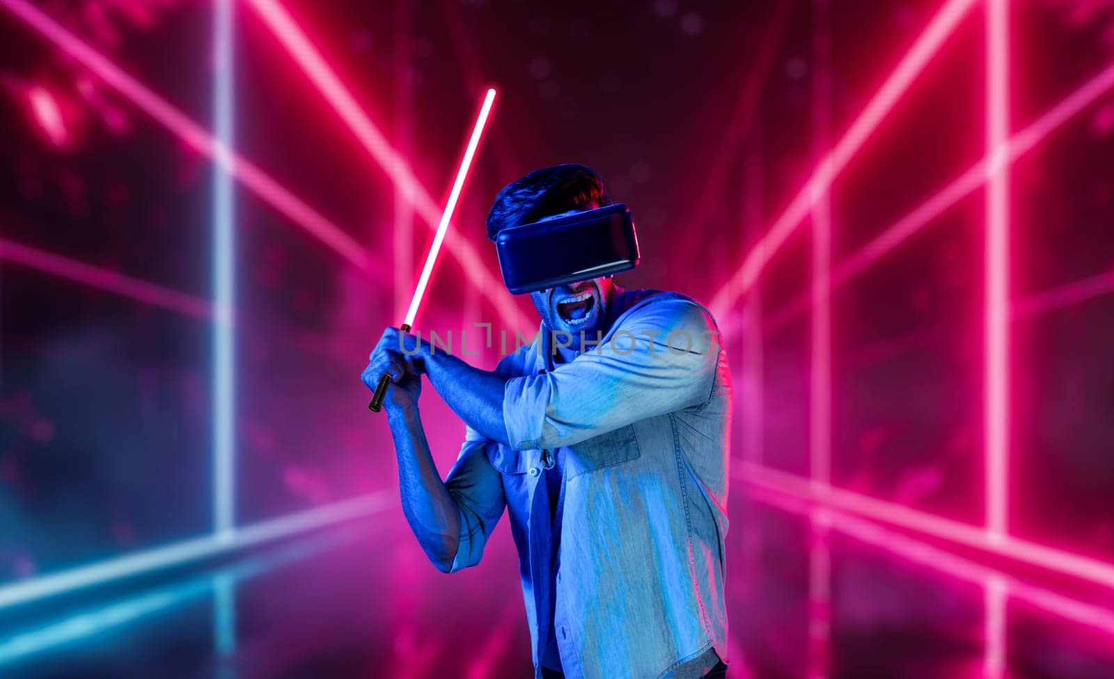 Gamer holding laser sword and playing action game while wearing VR glasses. Caucasian man using visual reality headset while standing and surrounding by neon castle. Innovation technology. Deviation.