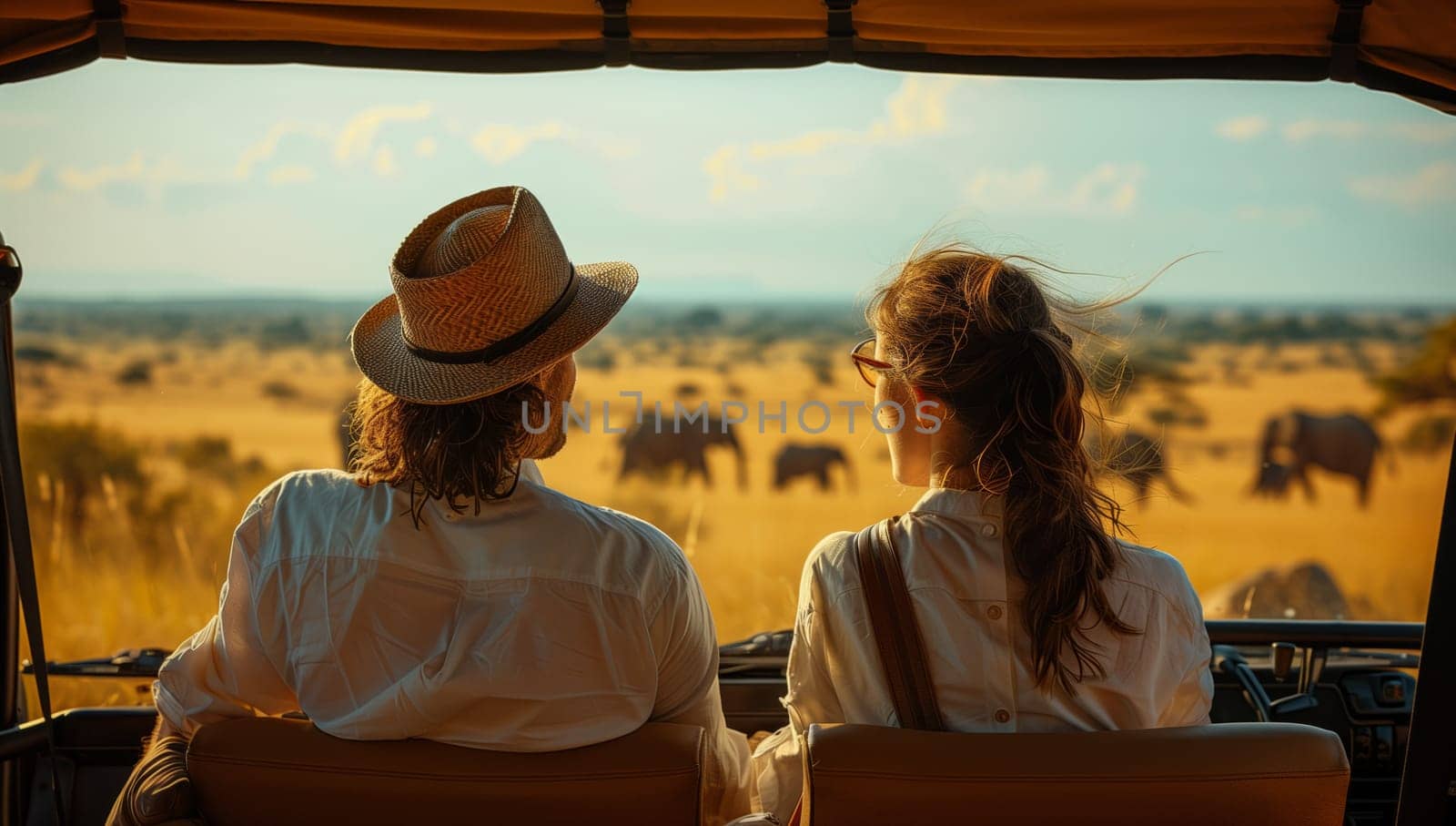 Woman and a man in sun hats are seated in the back of a jeep, admiring elephants in the landscape. They seem happy and are sharing a fun moment together under the sunny sky