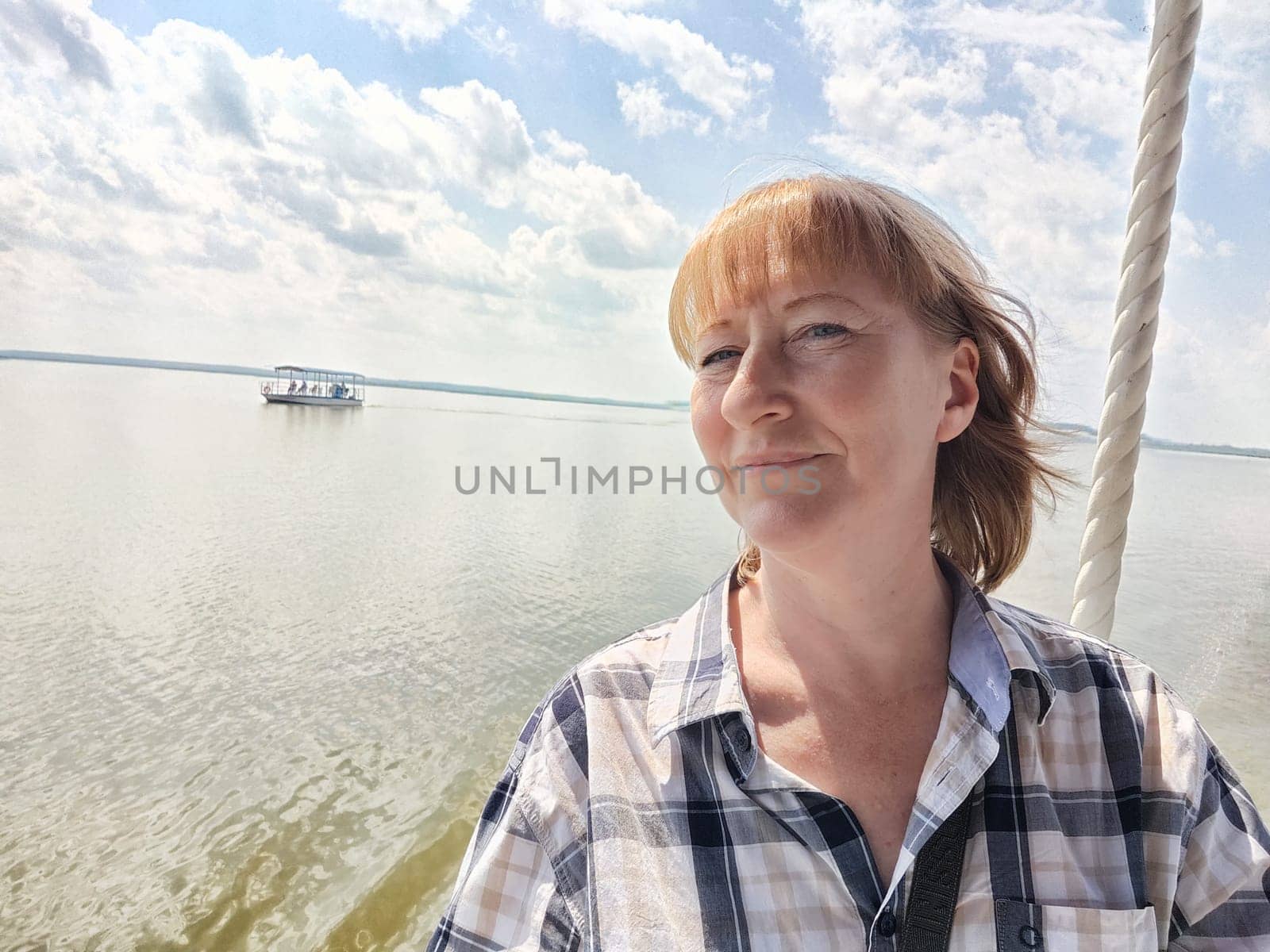 Happy cheerful middle aged woman taking selfie on nature outdoors and sun with water of lake or river on the background in summer day