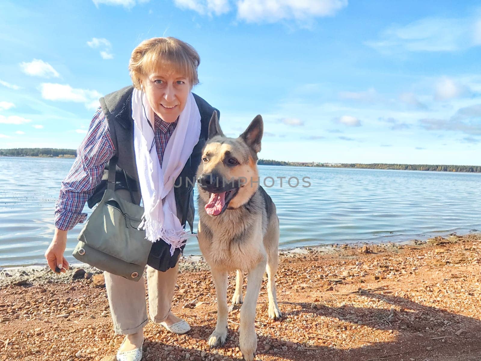 Adult girl with shepherd dog taking selfie near water of lake or river. Middle aged woman and big pet on nature. Friendship, love, fun, hugs