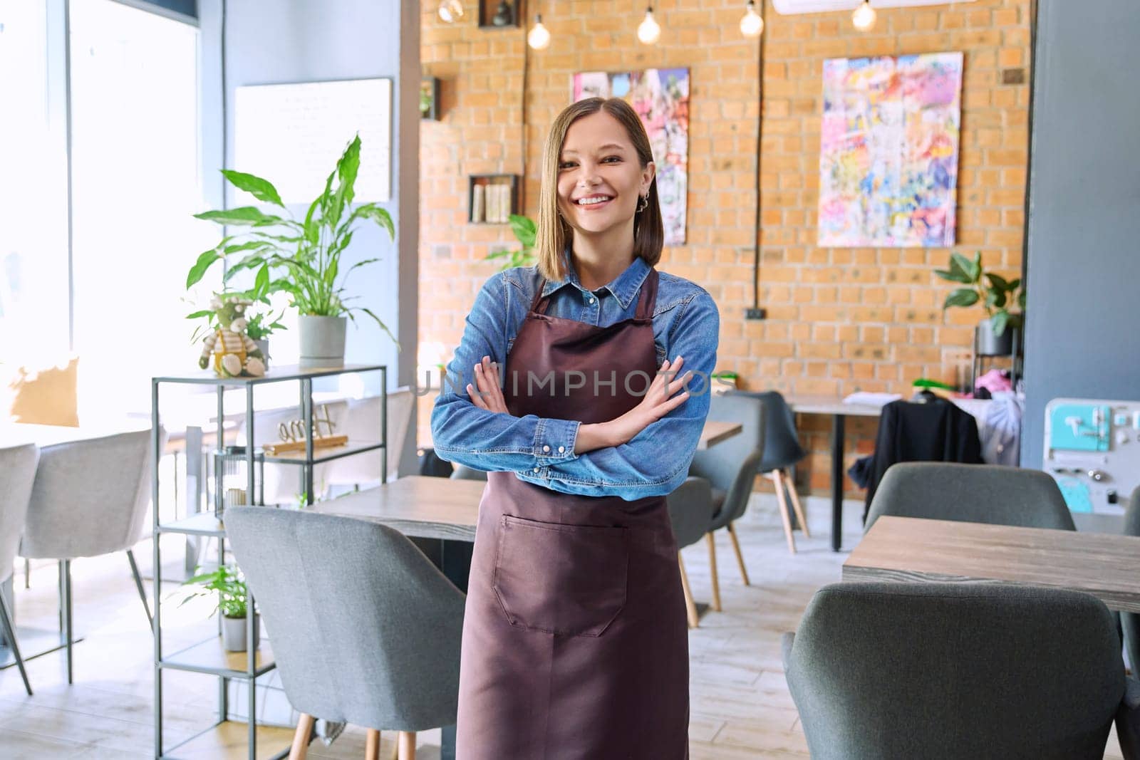 Confident successful young woman service worker owner in apron with crossed arms looking at camera in restaurant cafeteria coffee pastry shop interior. Small business staff occupation entrepreneur work