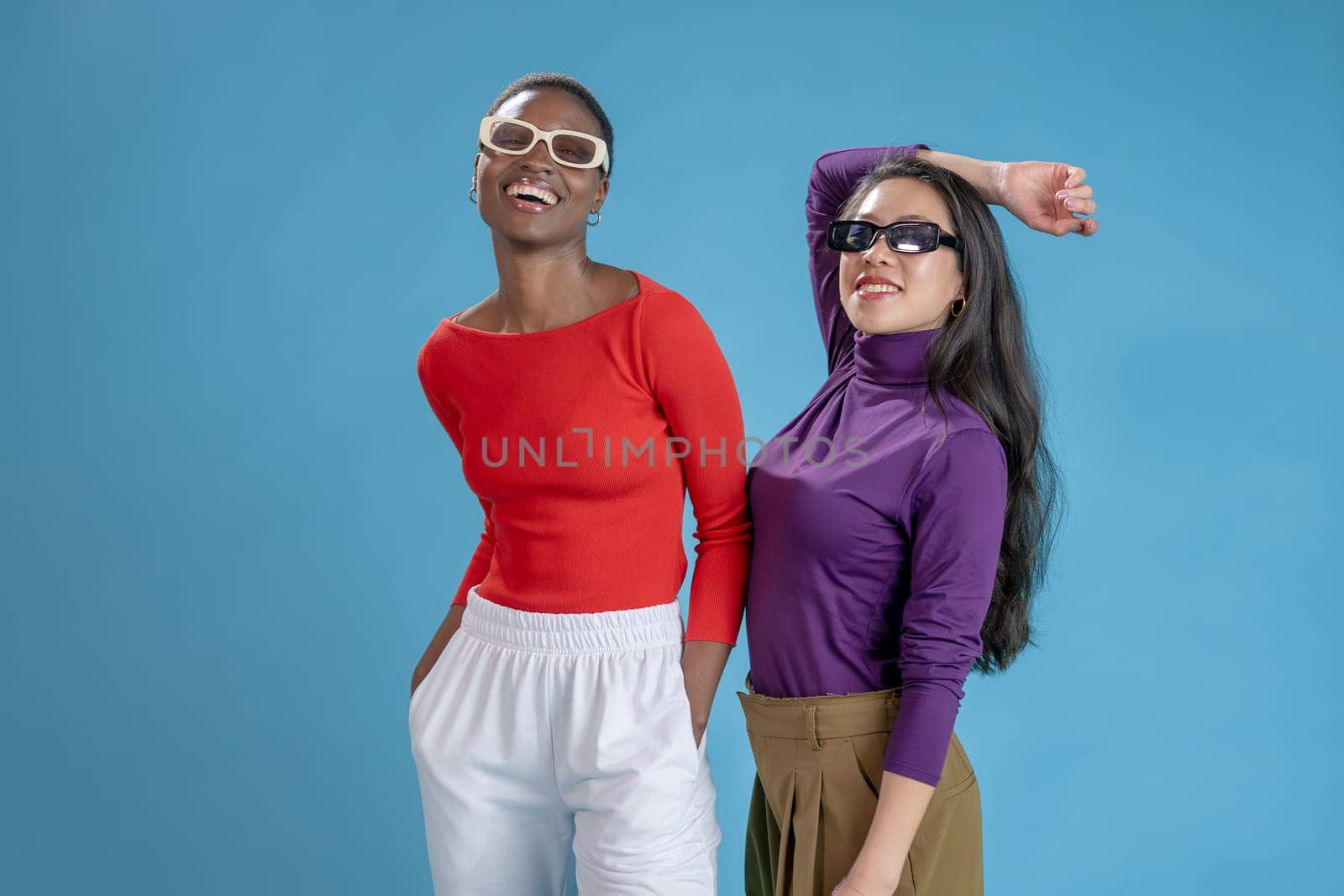 Two female fashion models with sunglasses posing smiling happy on a blue studio background by PaulCarr