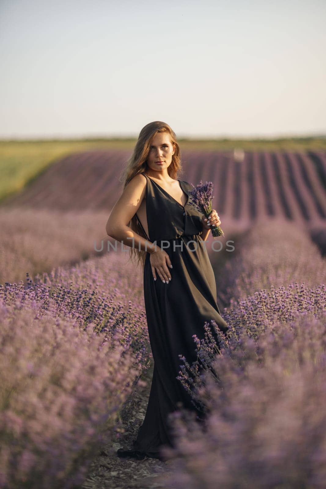 woman stands in a lavender field of purple flowers, holding a bouquet of flowers. The scene is serene and peaceful, with the woman taking a moment to enjoy the beauty of nature