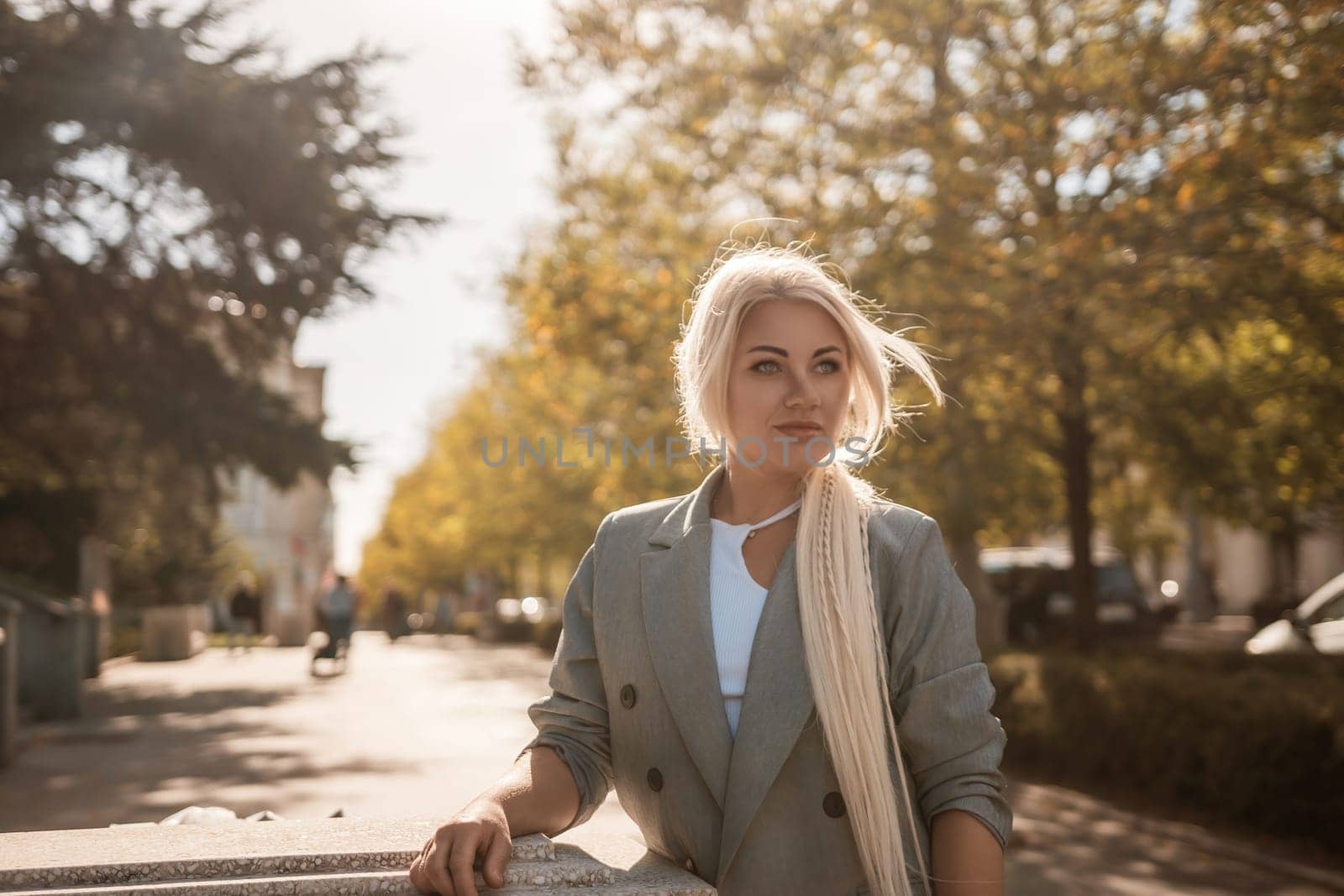 A blonde woman wearing a gray jacket and white shirt stands on a sidewalk. She is looking at the camera with a smile on her face. by Matiunina