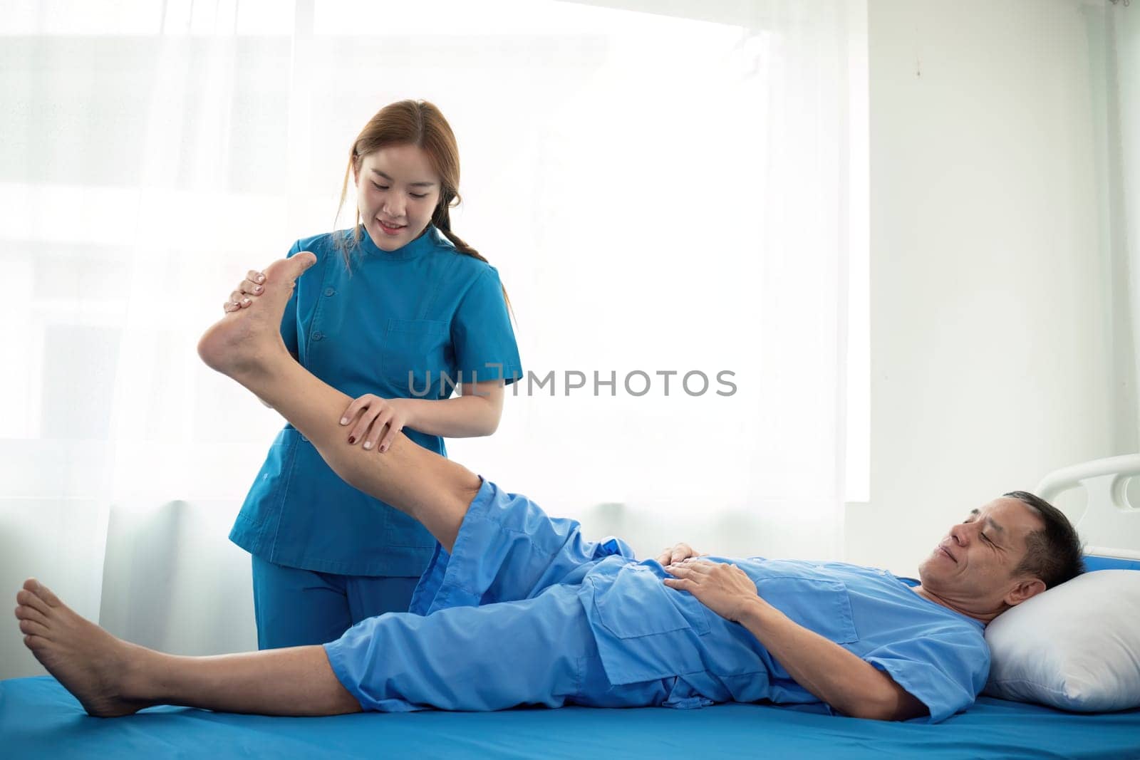 Physical therapist Asian woman, doing leg physiotherapy for elderly man to treat osteoarthritis and nerve pain in the leg to nursing at home and health care concept by nateemee