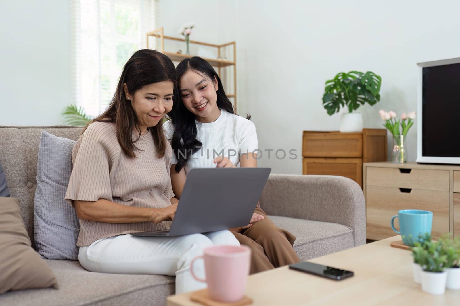 Mother and adult daughter sitting on the sofa together, mother and daughter using laptop to surf website together by itchaznong