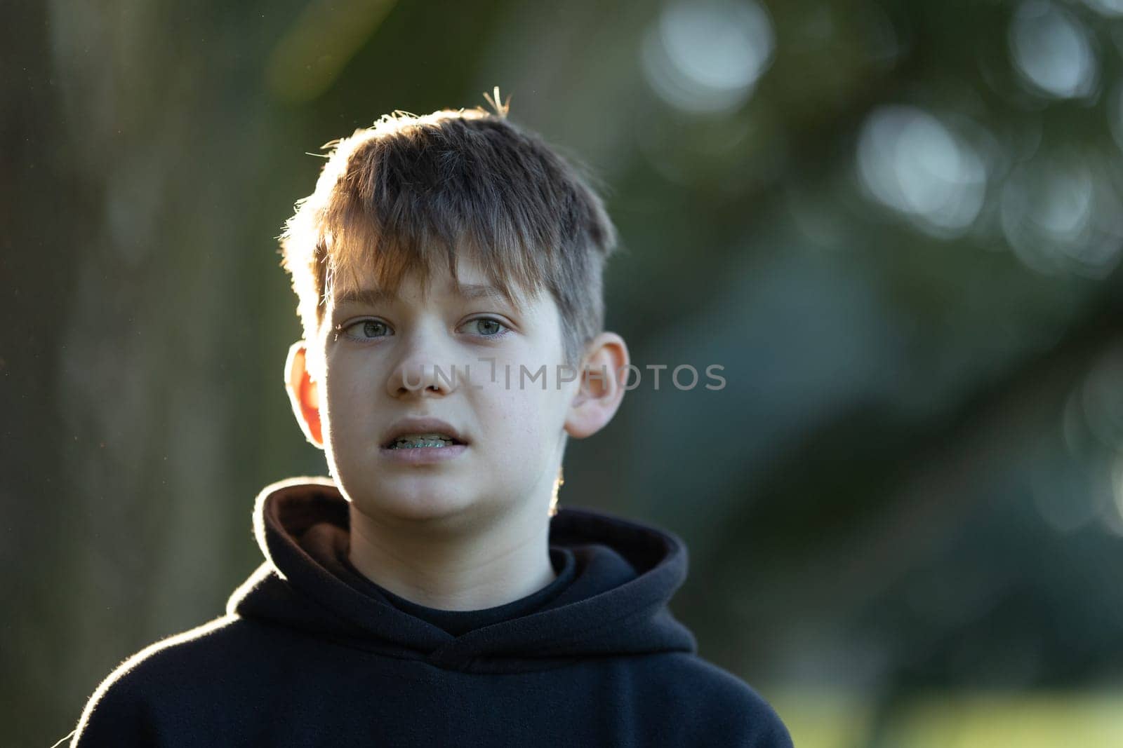 A boy with a black hoodie and a blue shirt is standing in the sun. He has a serious expression on his face