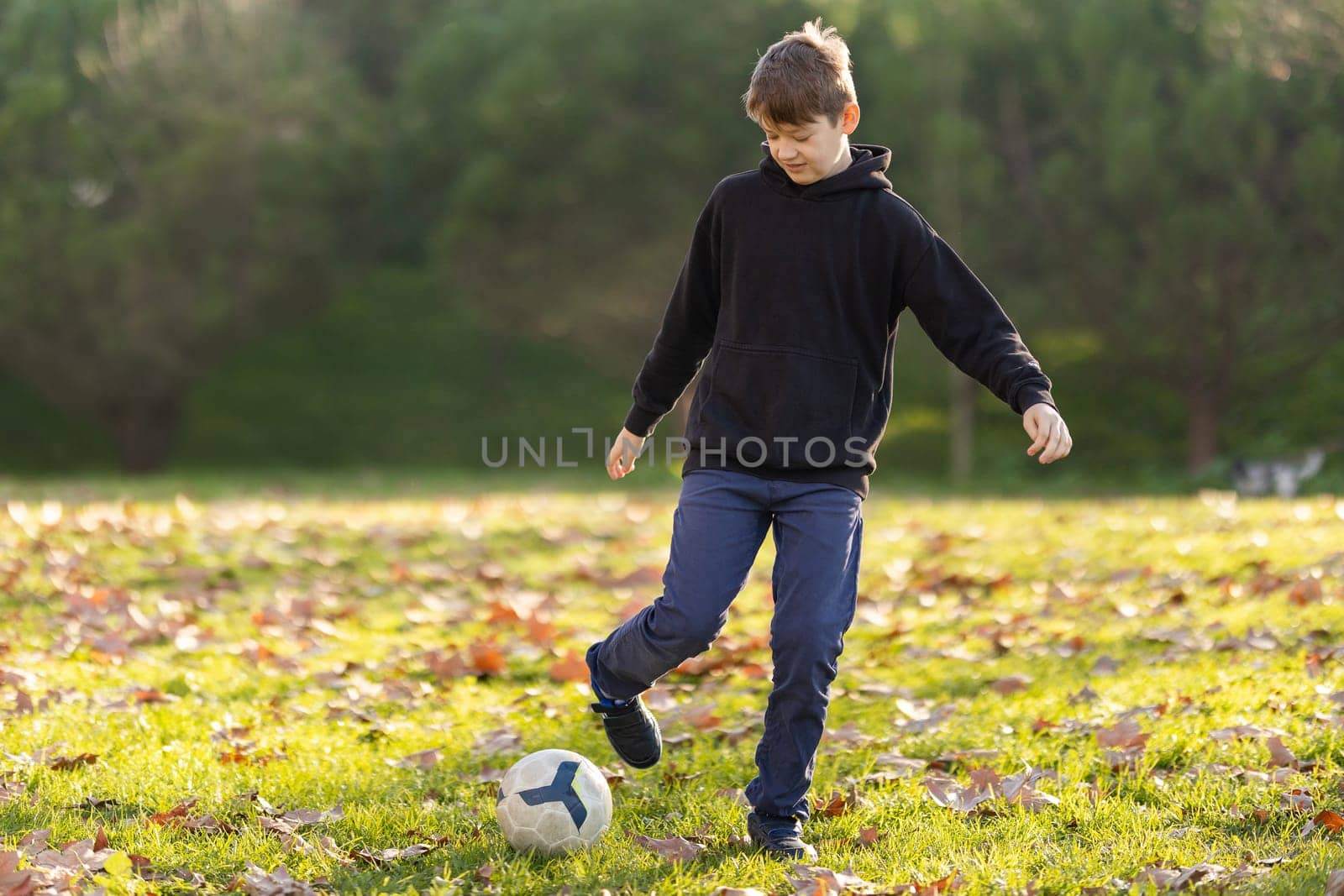 A boy kicks a soccer ball in a park. The boy is wearing a black hoodie and blue jeans