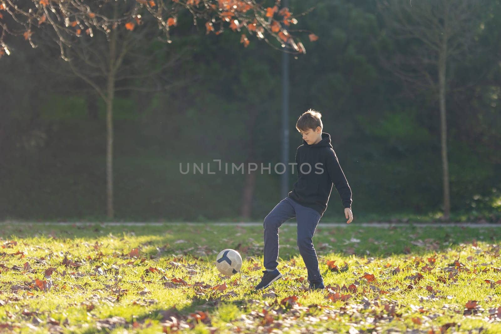 A boy is kicking a soccer ball in a park. The boy is wearing a black hoodie and blue jeans. The park is filled with trees and leaves, creating a peaceful and natural atmosphere