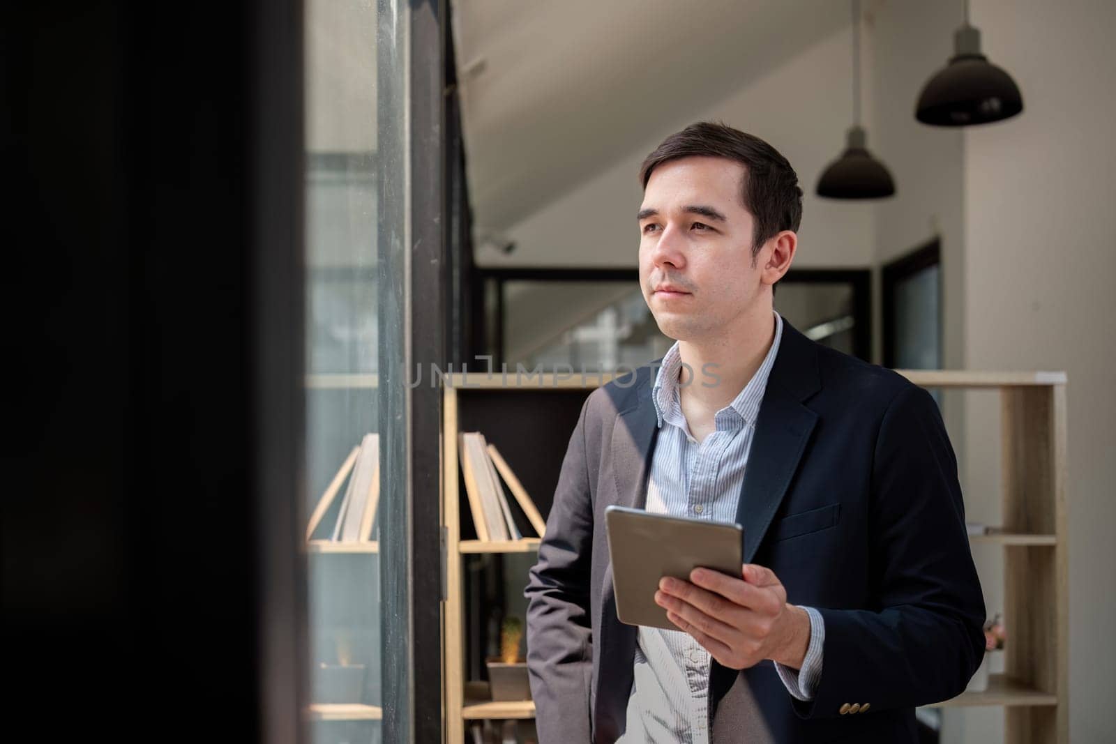 Happy young businessman using online app on tablet for job communication, looking out the window at office.