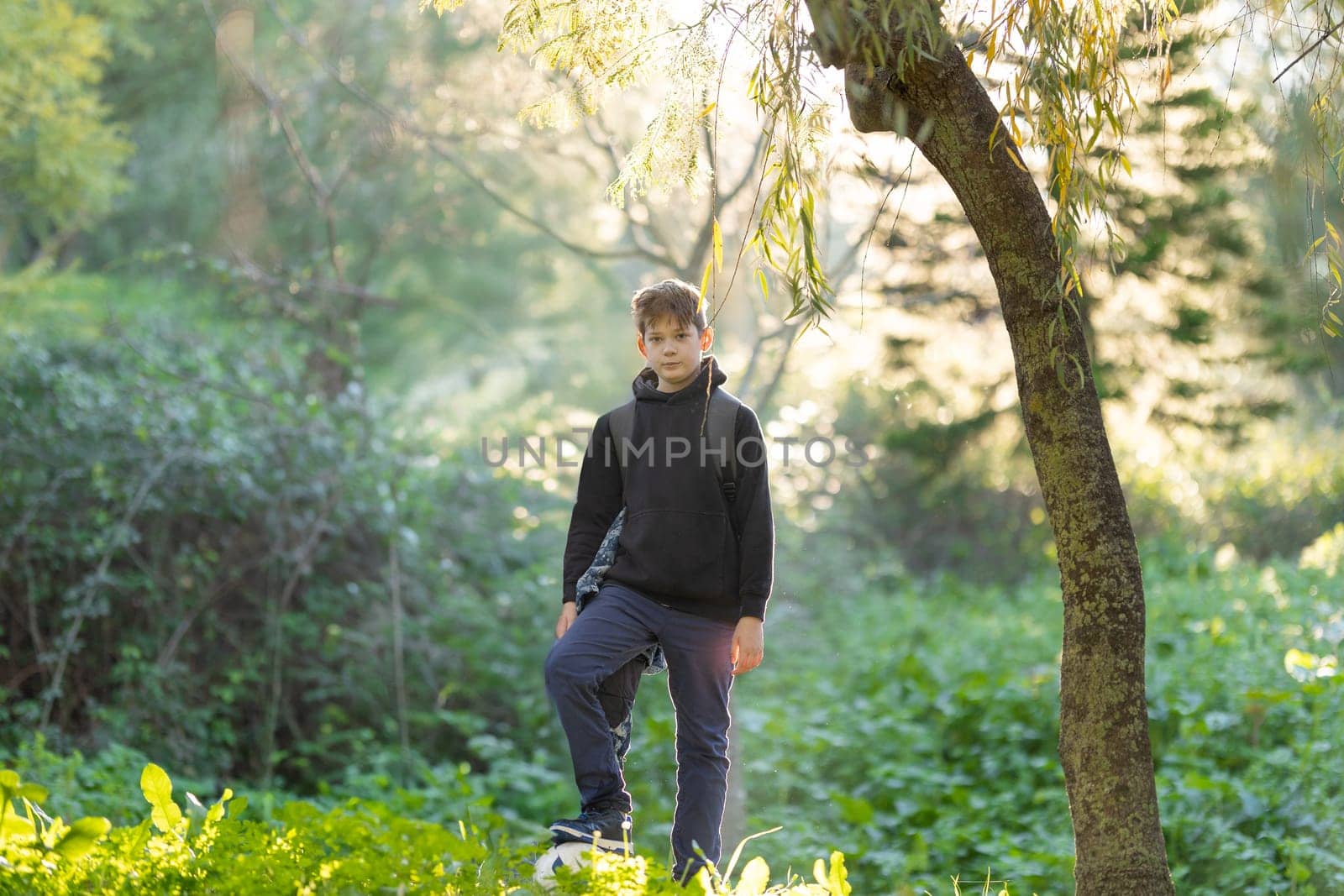 A boy is standing in a forest with a backpack on. The boy is wearing a black hoodie and blue pants