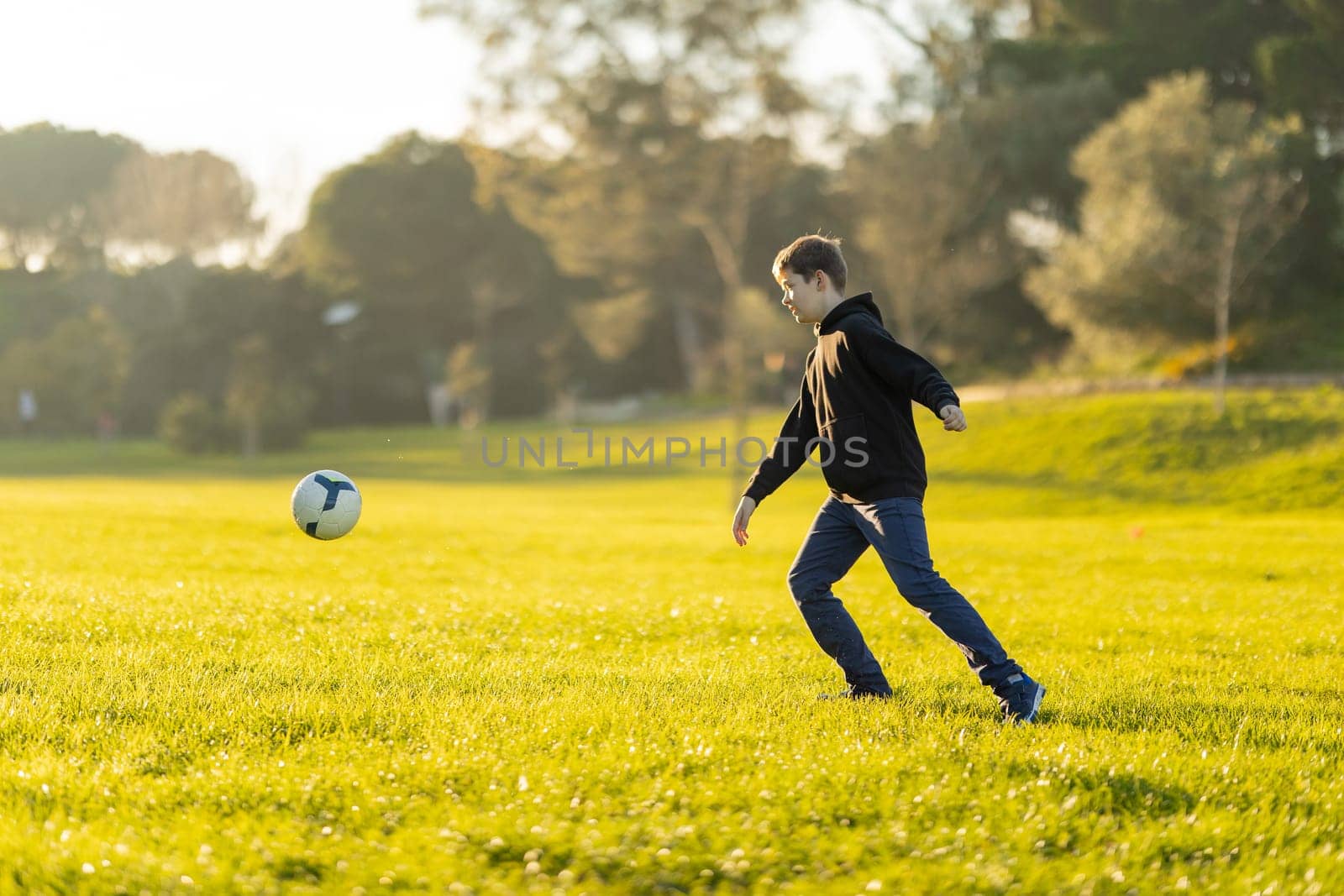 A boy kicks a soccer ball in a field. The boy is wearing a black hoodie and blue jeans