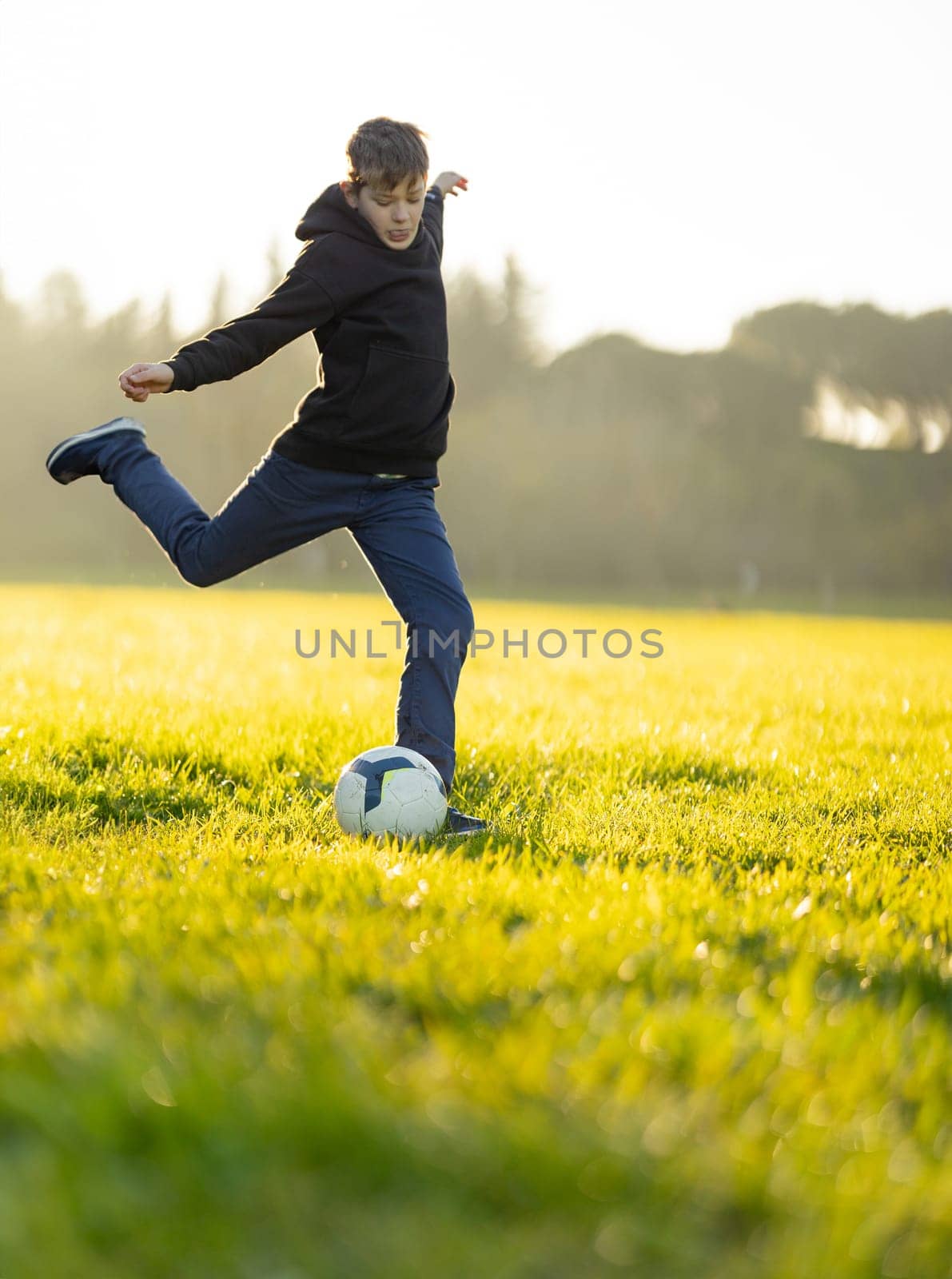 A boy kicks a soccer ball in a field. The boy is wearing a black hoodie and blue jeans