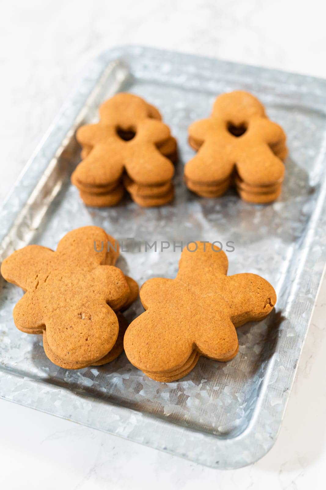 Gingerbread cookies, including a gingerbread man with a heart-shaped cutout, rest on a rustic metal tray against a marble countertop.