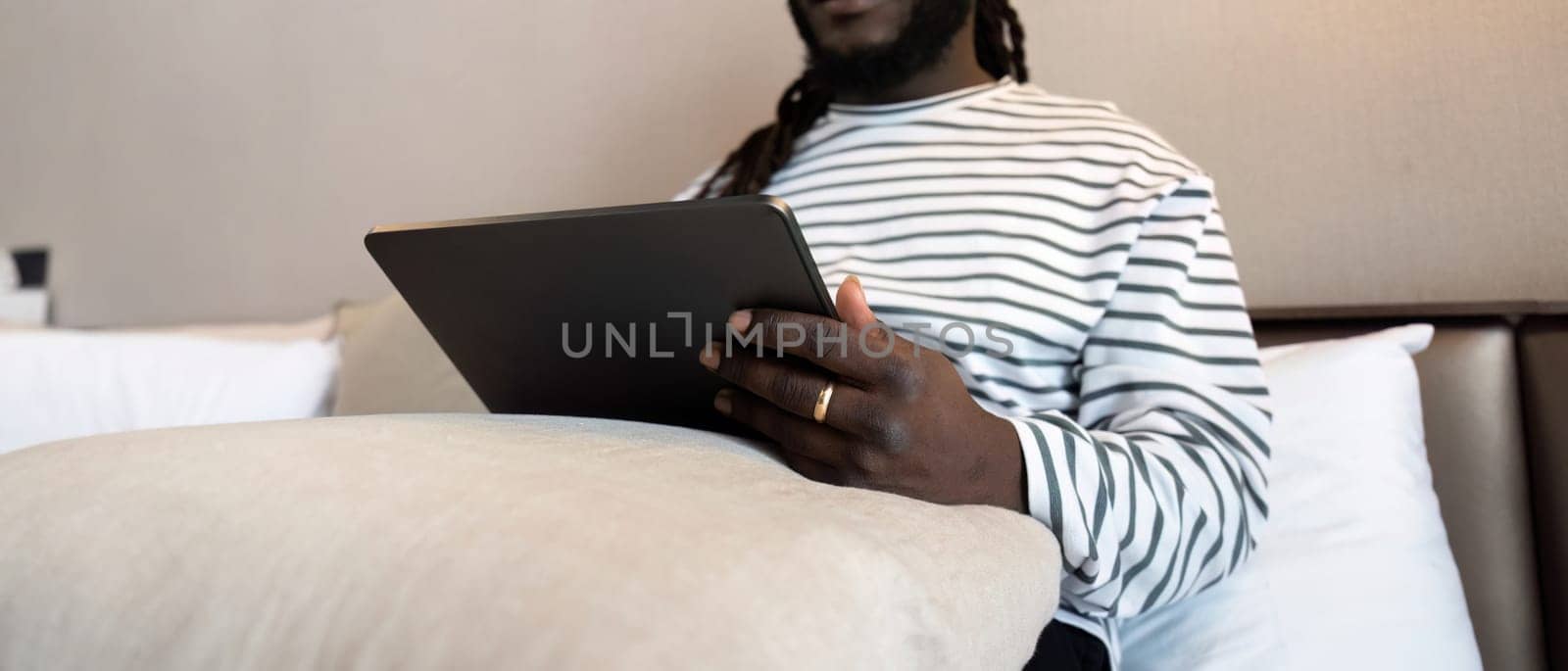 Smiling young African American man using digital tablet remote job sitting on the bed at home. concept online technology communication.