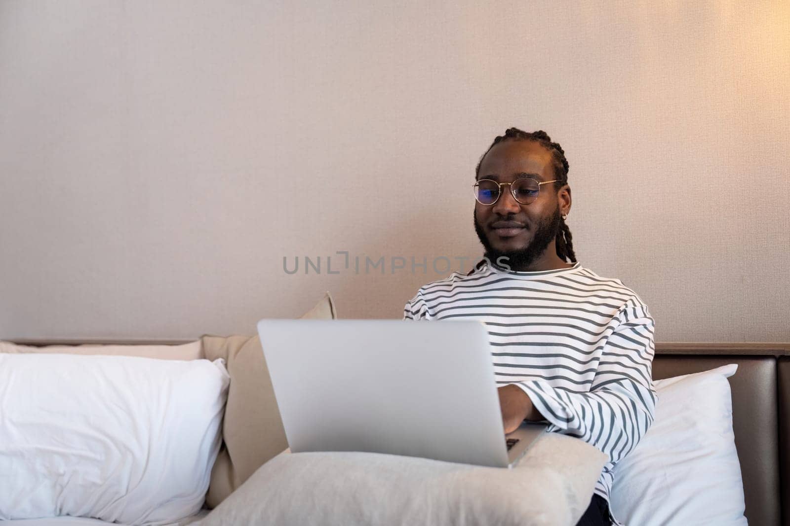 Smiling young African American man using laptop remote job sitting on the bed at home. concept online technology communication by nateemee