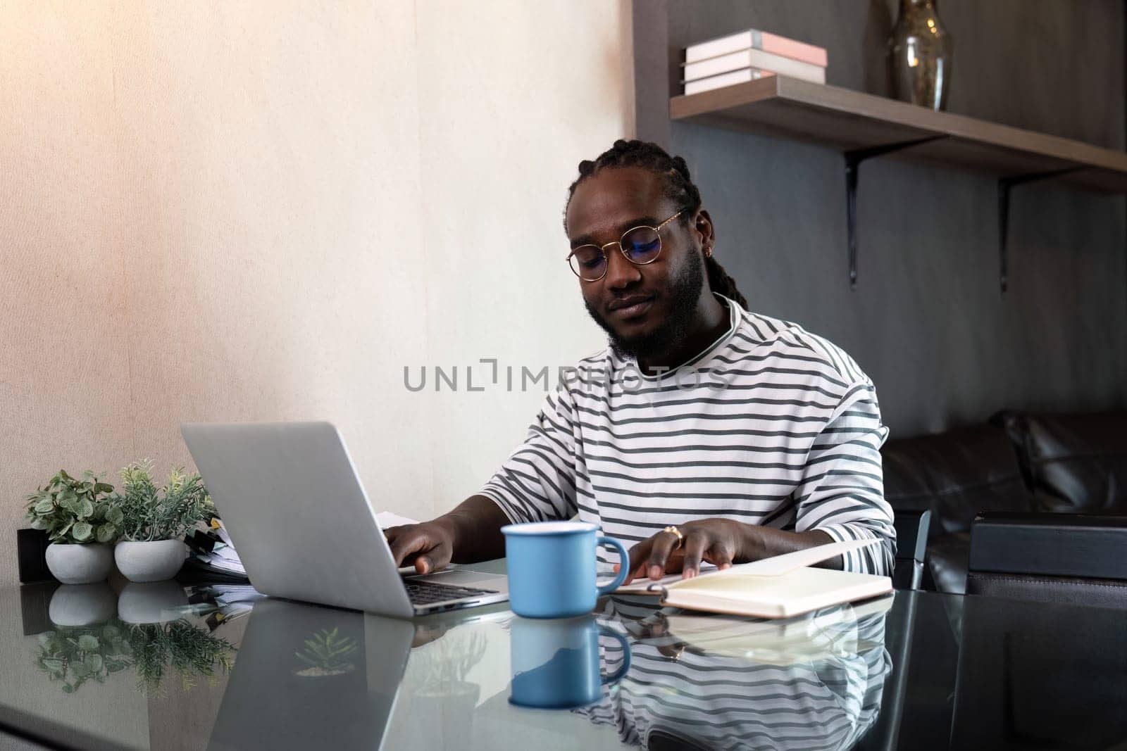 African American man working with laptop computer remote while sitting at glass table in living room. Black guy do freelance work at home office by nateemee