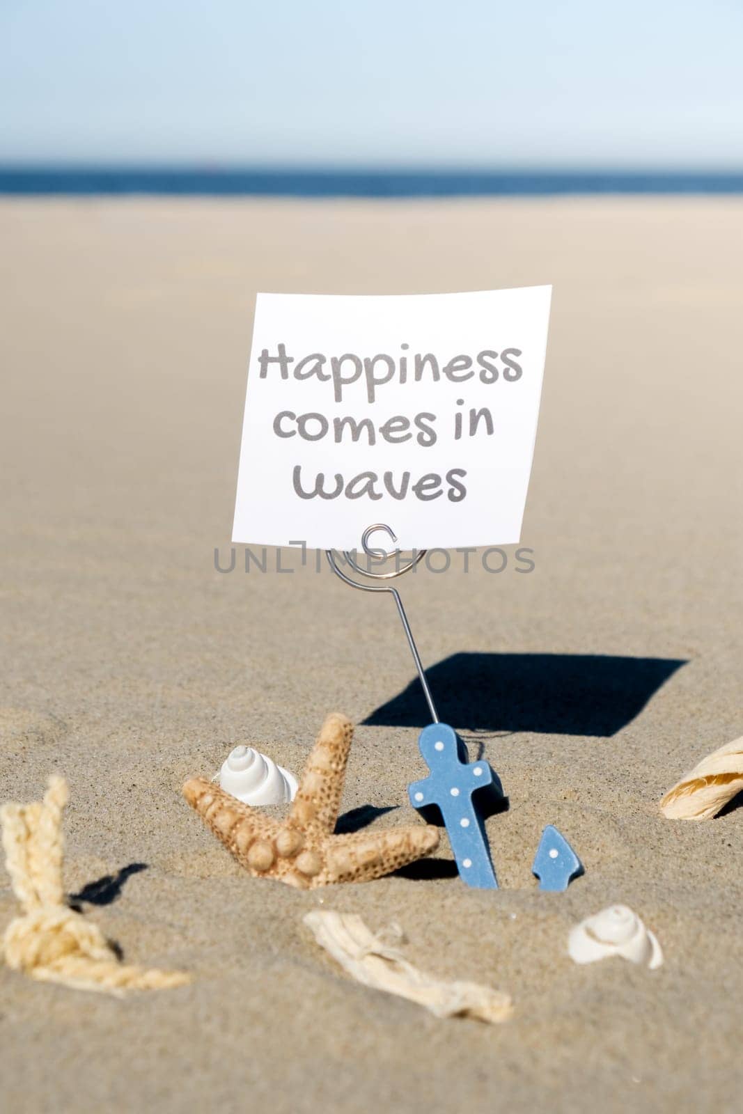 HAPPINESS COMES IN WAVES text on paper greeting card in anchor paper holder and starfish seashell summer vacation decor. Sandy beach sun coast. Holiday concept postcard. Getting away Travel by anna_stasiia