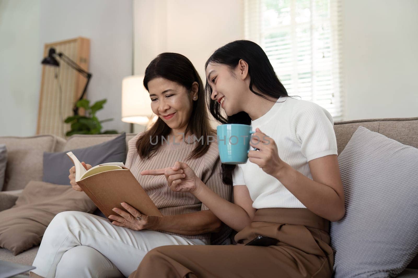 Happy senior mother with adult daughter sitting on couch and holding cups with coffee or tea at home. Enjoy family concept by nateemee