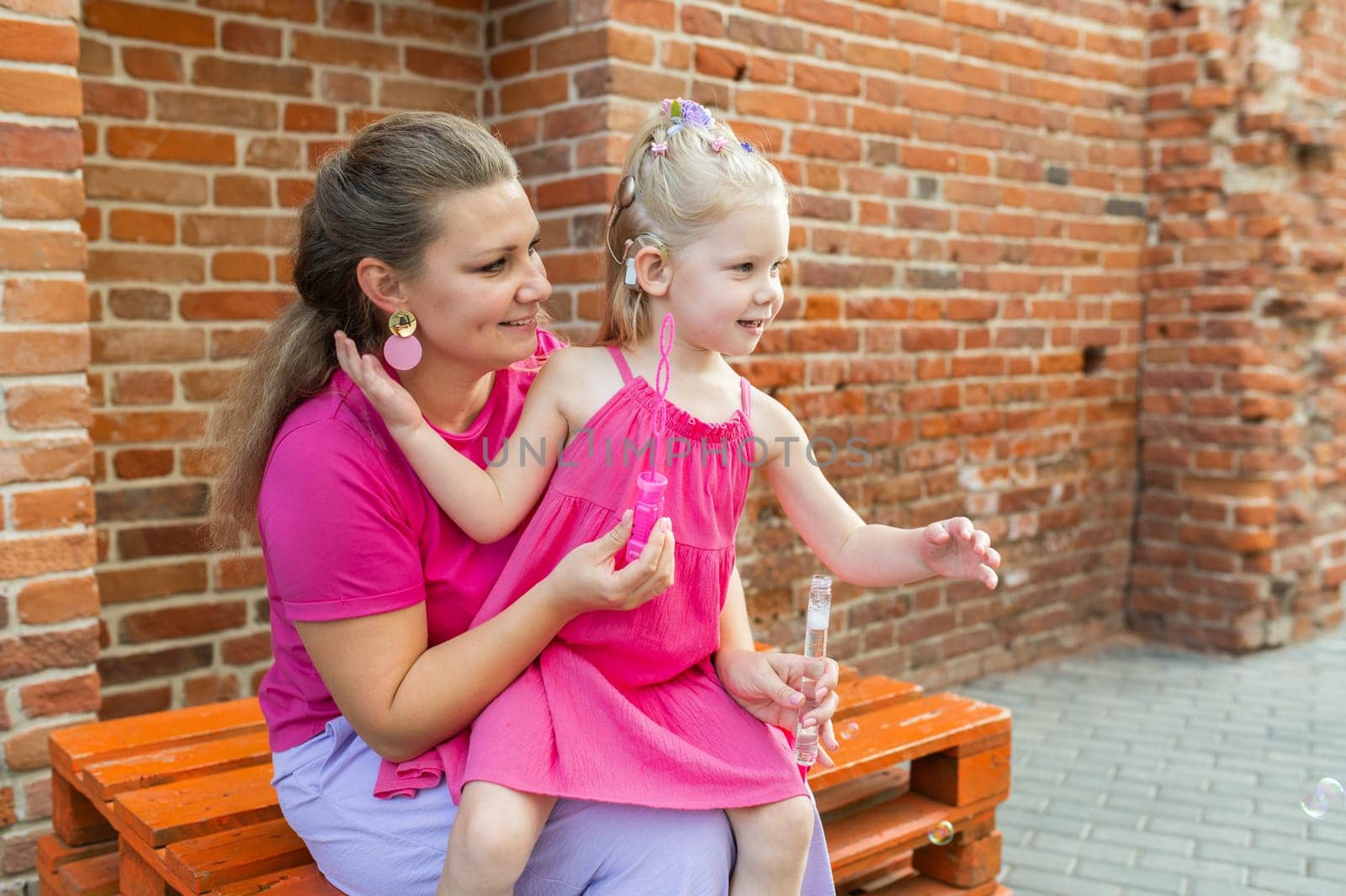 Deaf child with cochlear implant for hearing audio and aid for impairment having fun and laughs with mother outdoor in summer. Sound fitting device to help with communication listening and interaction.