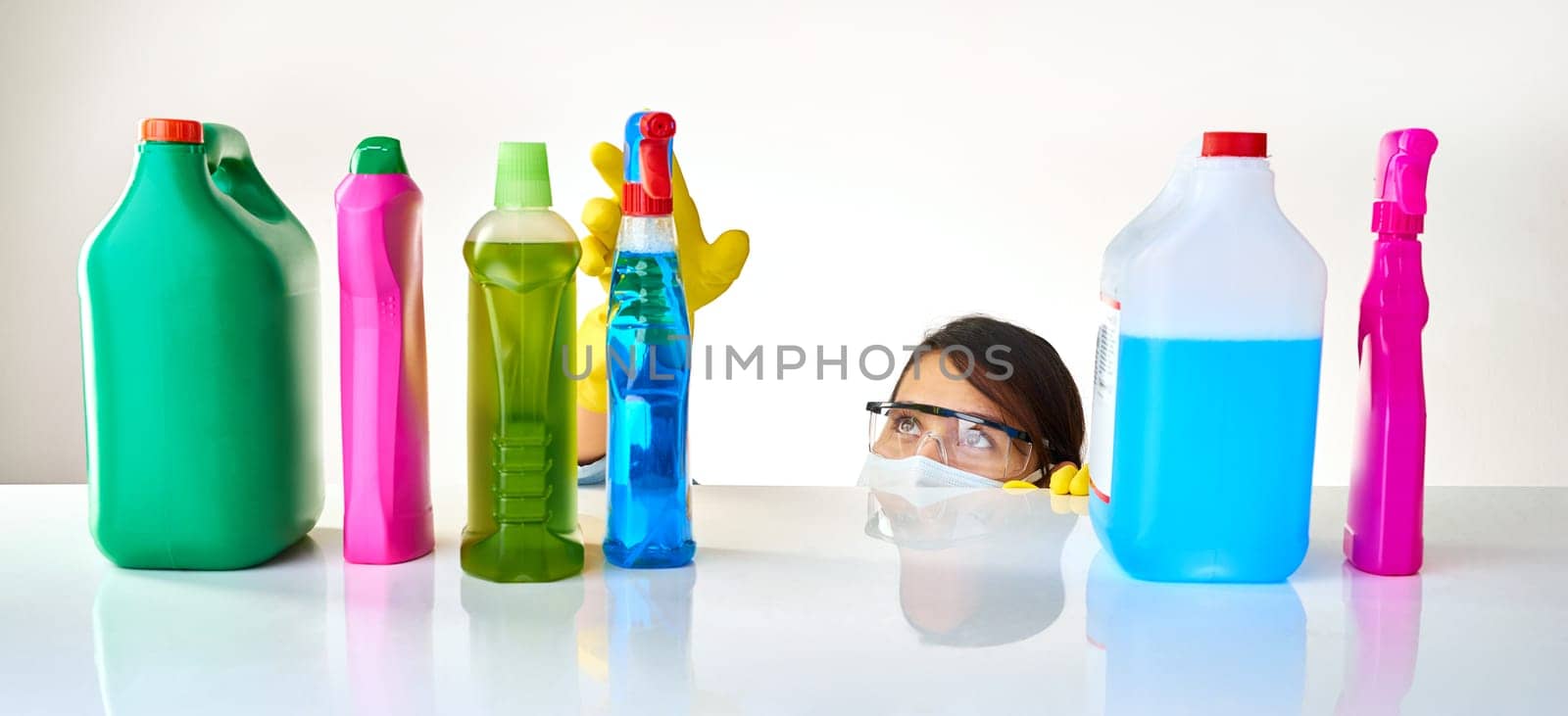 Detergent, products and woman with mask for cleaning or glasses for protection, safety and soap at home. Housekeeping, cleaner or female person with bottles, liquid container or spray on table.