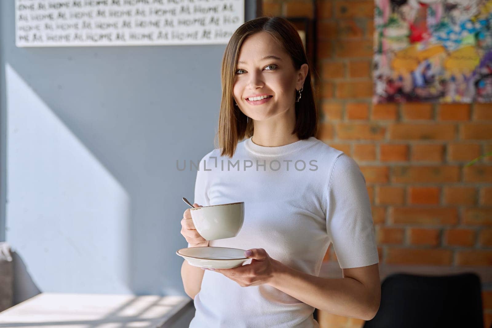 Young beautiful happy woman holding cup of coffee tea with saucer looking at camera, gray wall of cafe coffee shop cafeteria copy space for text. Coffee business work services youth lifestyle leisure