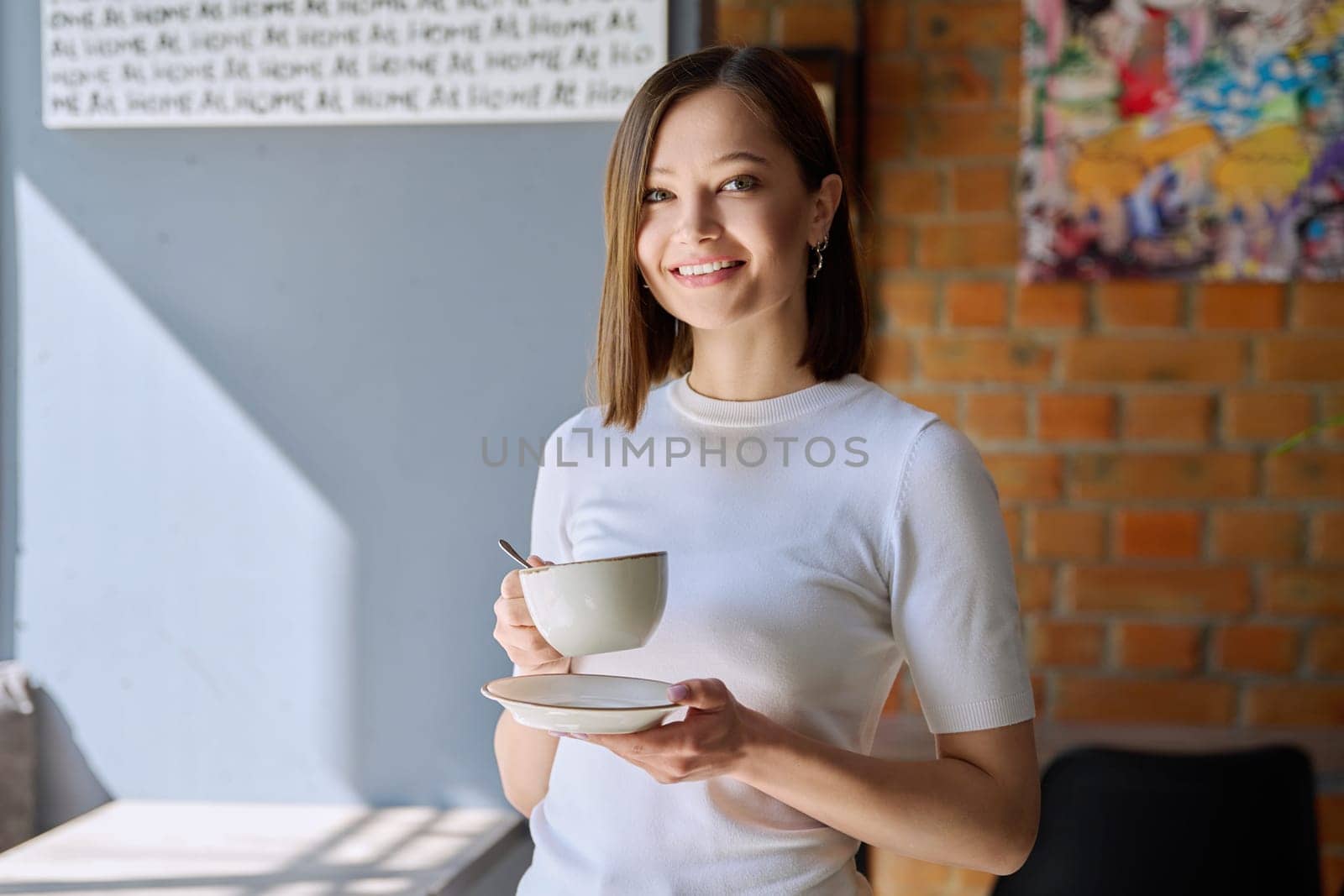 Young beautiful woman holding cup of coffee, looking out window, gray wall copy space by VH-studio