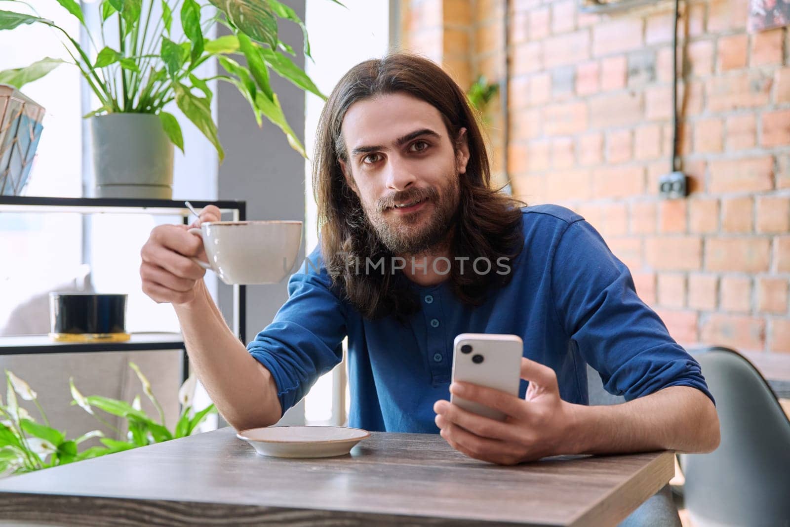 Young handsome man using smartphone, drinking cup of coffee, sitting in cafe by VH-studio