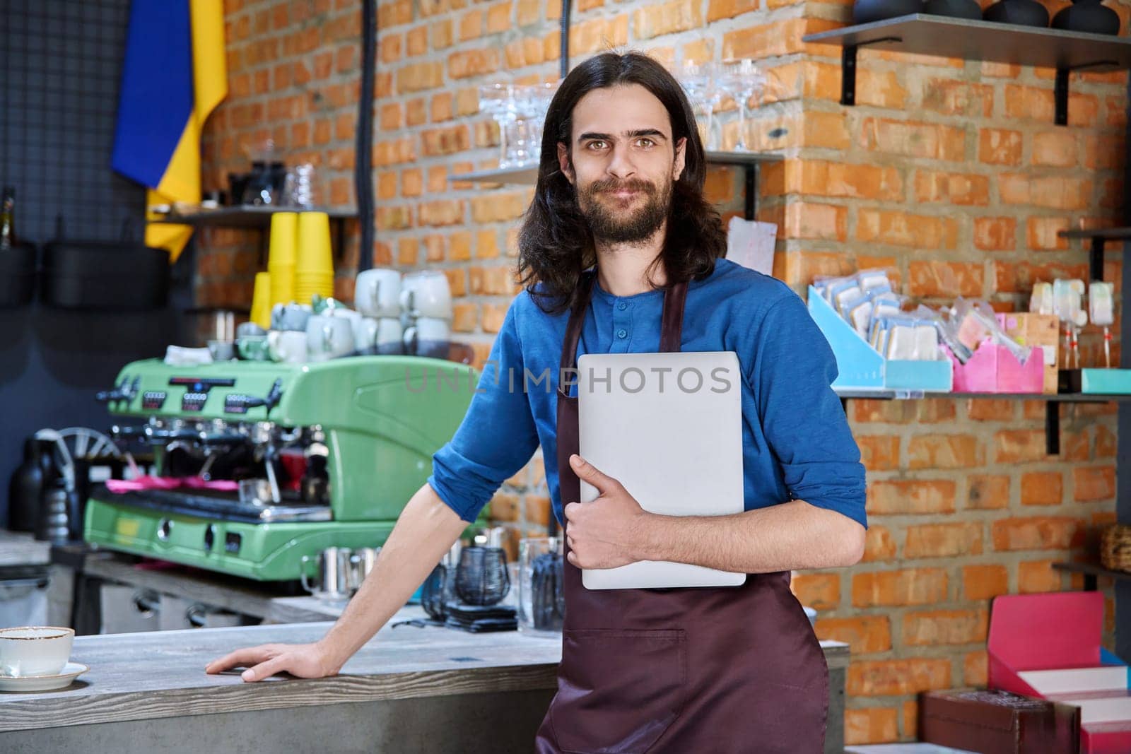 Young man in apron, food service worker, small business owner holding laptop by VH-studio