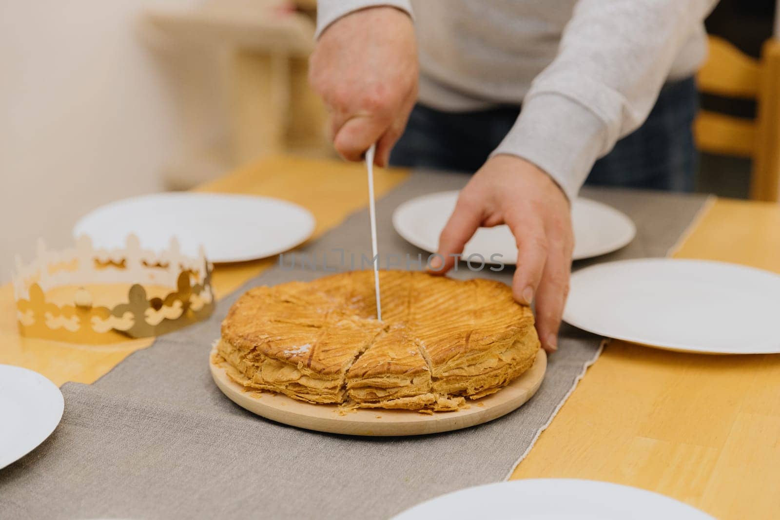 One Caucasian young blurred man cutting a king's galette in a plate on a wooden board using a knife while standing at the table, close-up side view with selective focus.
