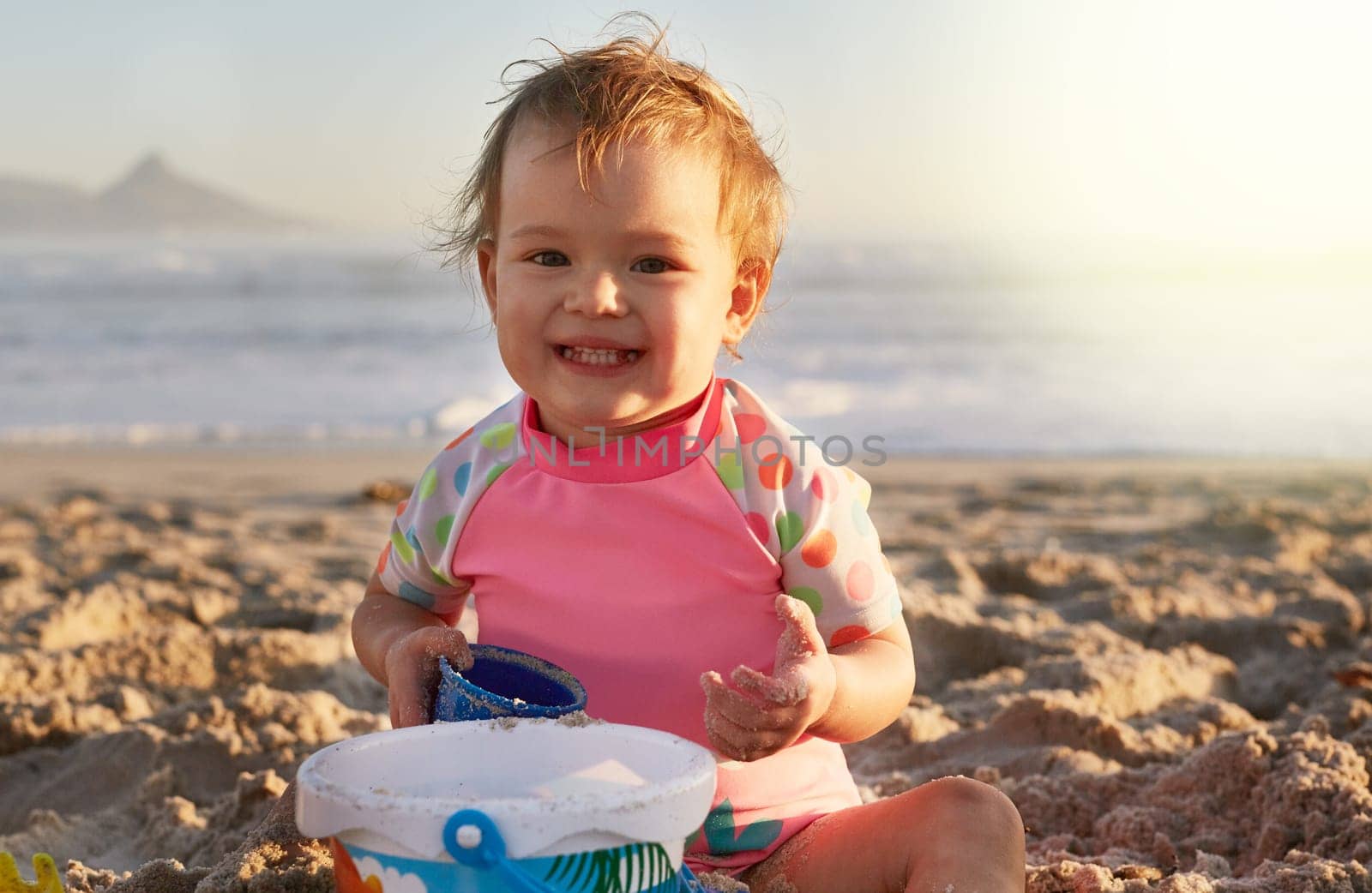Girl, baby and playing in sand on beach with portrait, toy bucket and happy with swimwear in summer. Child, toddler and smile with relax on holiday, vacation and fun with sunlight by ocean in nature.