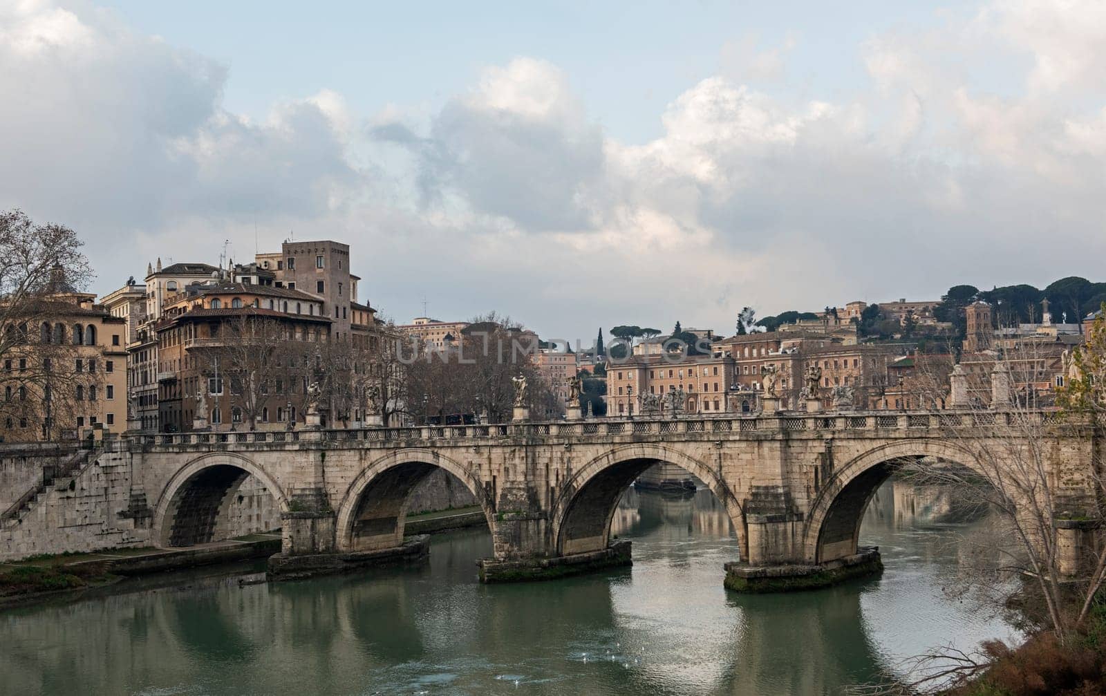 Old stone arched road bridge over large river Tiber in Rome Europen city centre with overcast cloudy sky