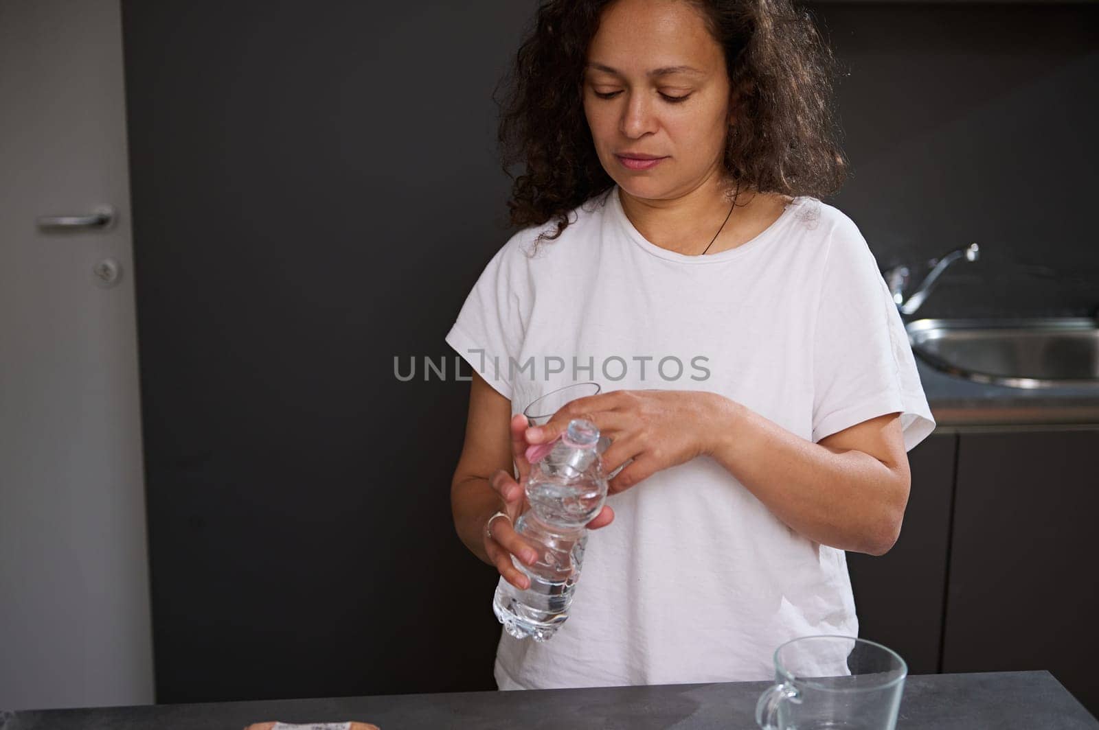 Portrait of multi ethnic sleepy woman in white pajamas, holding bottle of pure water, starting her day with sip of refreshing water for healthy diet or detox. People. Healthy active lifestyle concept by artgf