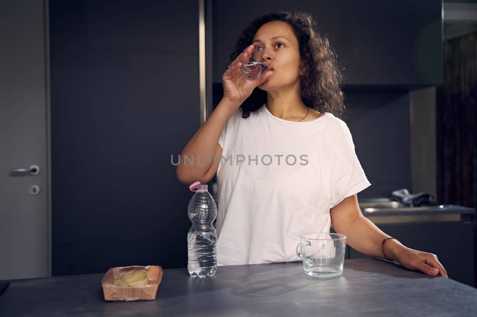 Beautiful multi ethnic curly haired young adult woman in white pajamas, standing at kitchen table and drinking pure water from glass while waking up in the morning. Healthy lifestyle concept. by artgf